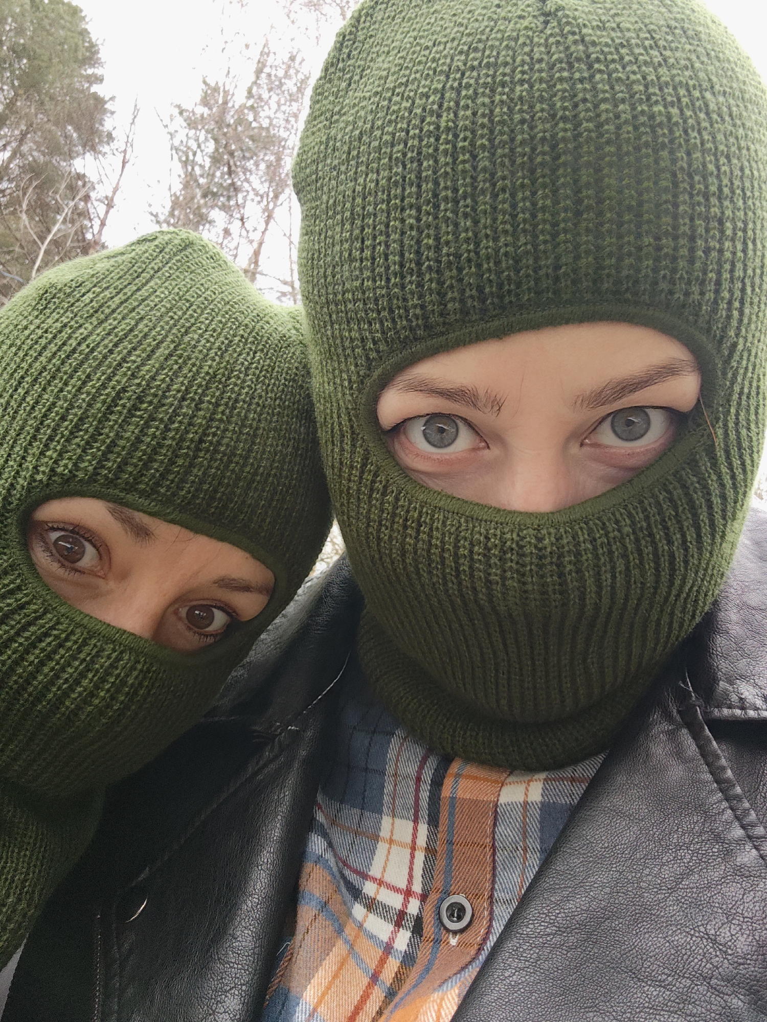 Two individuals wearing green balaclavas are closely positioned, suggesting a possible selfie. The person on the right wears a black leather jacket over a plaid shirt with a button visible. The background shows bare trees, suggesting an outdoor setting. Without further information, location in the city of Chaikovsky, Russia is not confirmed.