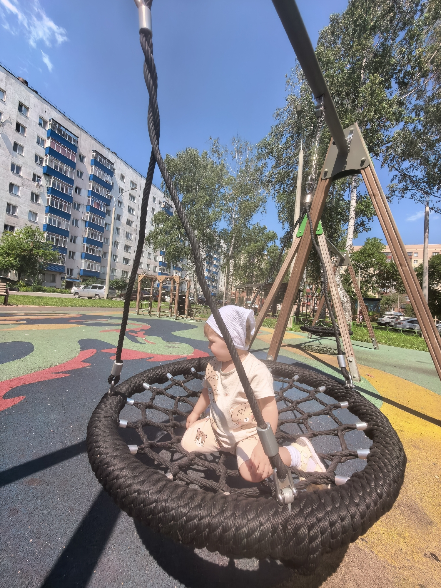 In Sterlitamak, Russia, a young child is seated on a black, circular net swing at a playground. The child is wearing a light-colored outfit with animal prints and a white headscarf. The playground surface is a multi-colored rubberized material. In the background, a tall, white apartment building with blue balconies is visible, along with trees and other playground equipment. The sky is clear and blue, indicating a sunny day.