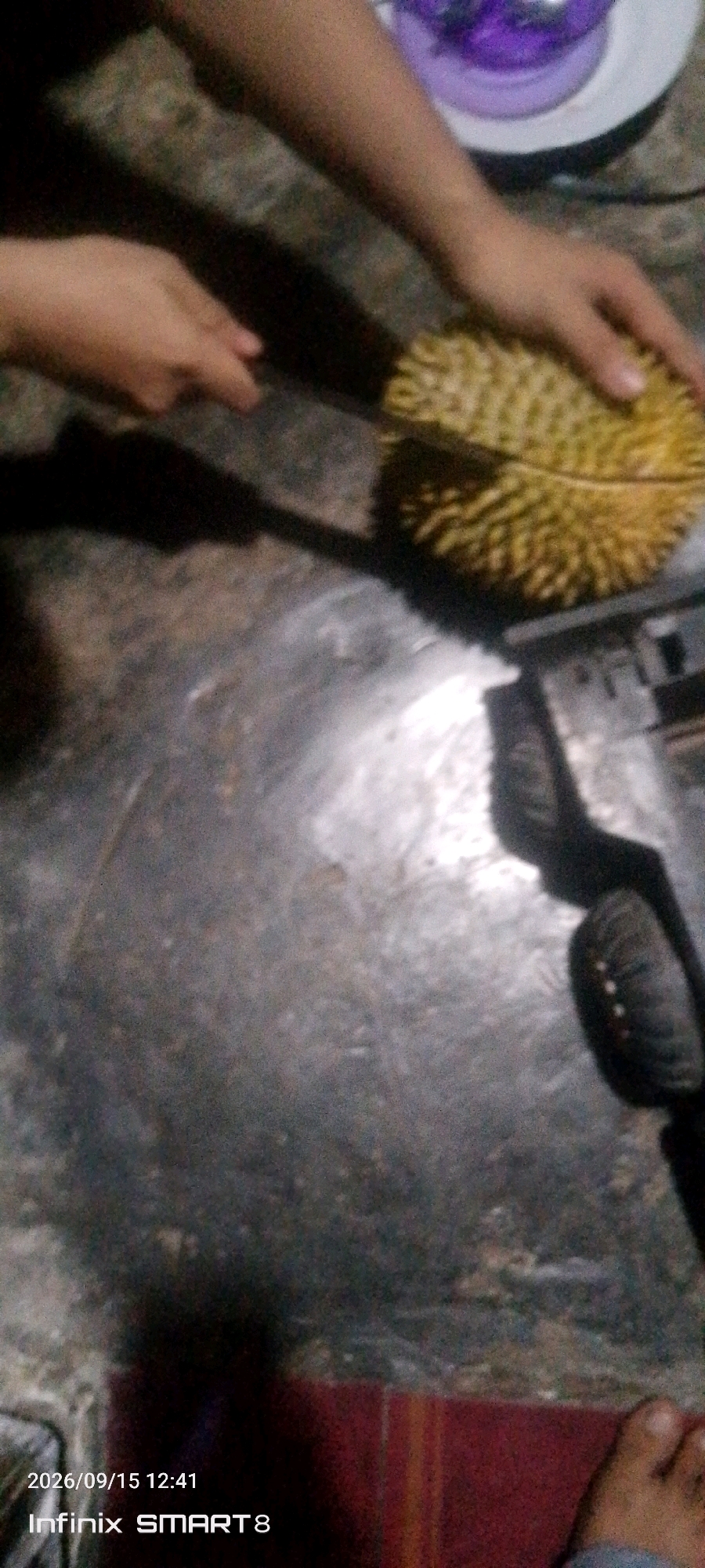 In the city of Serang, Indonesia, an individual is in the process of cutting a durian fruit. The scene is dimly lit, suggesting it might be indoors or at night. The durian, a large spiky fruit, is partially cut, revealing its segments. A knife is being used by the individual to divide the fruit. The surface beneath the fruit appears to be a floor made of concrete or tile, with visible texture and some debris. Shadows are cast on the floor, indicating a light source from above and to the side. There are other objects in the background, out of focus, including what appears to be a purple object and possibly a hand on the left side of the frame, also out of focus. The bottom right corner of the image shows a person's foot, suggesting the photographer's presence. The timestamp in the lower left corner indicates the photo was taken on September 15, 2026, at 12:41, with the device identified as an Infinix SMART 8.