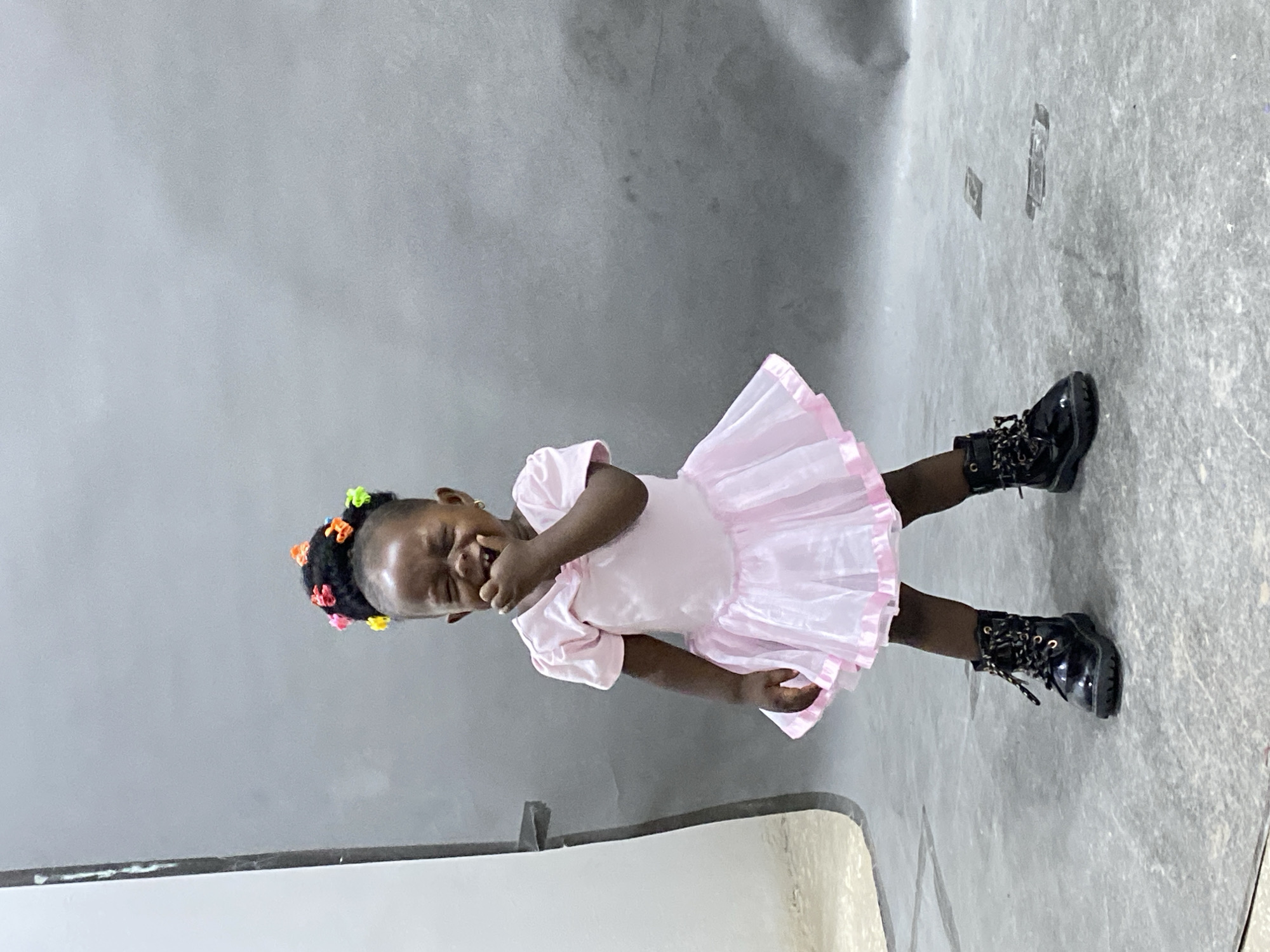 A young girl wearing a pink dress and black boots stands in a studio. The background is a gray wall and floor. The girl is covering her mouth with her hand. She has colorful hair clips in her hair. The location is Gura Top, Nigeria, but the studio could be anywhere.