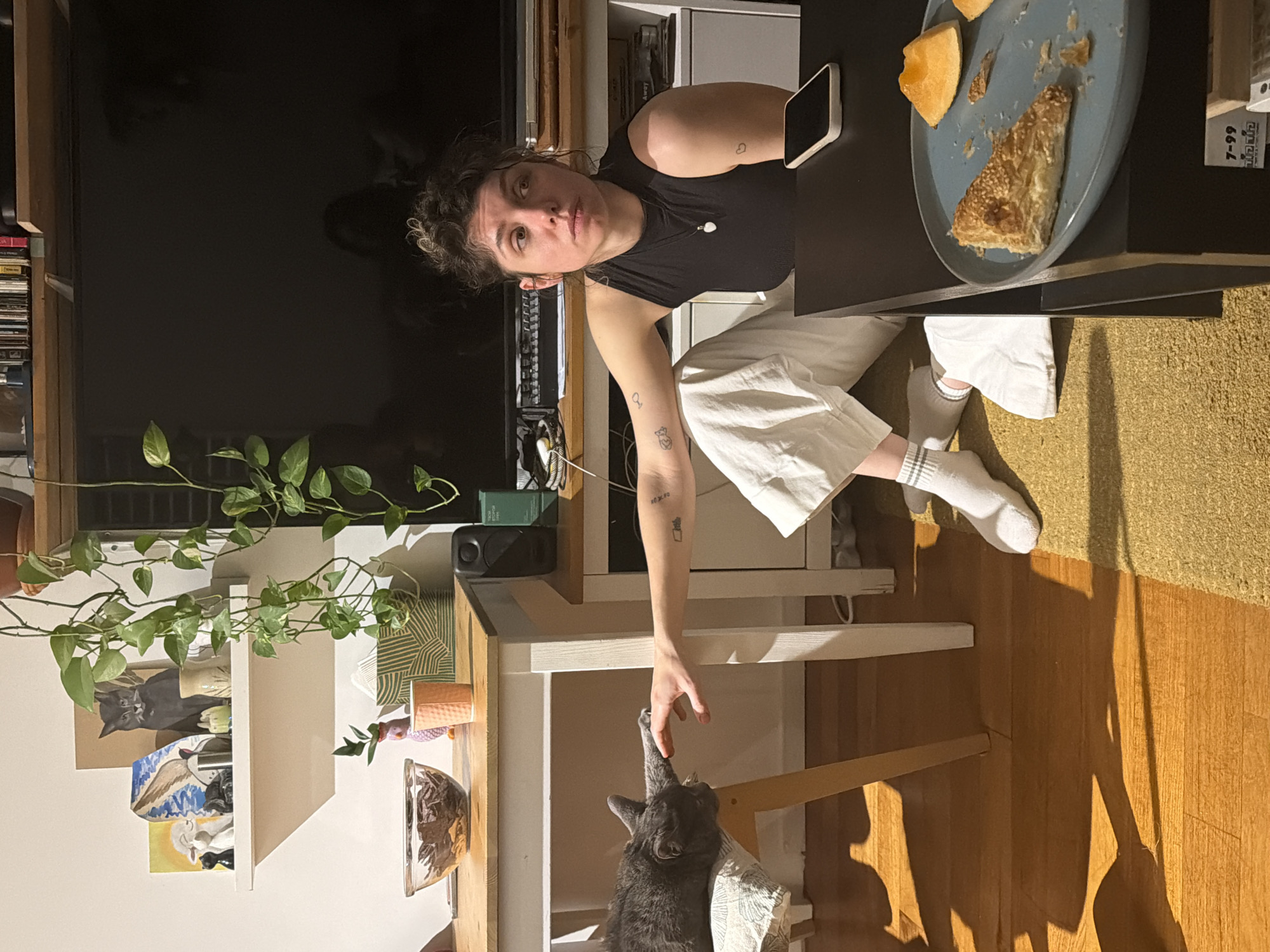 This indoor scene captures a woman interacting with a cat in what appears to be a home in Tel-Aviv, Israel, likely in the evening or at night.
The primary subject is a woman with dark, curly hair, wearing a black tank top and white, loose-fitting pants. She is seated at a dark table, looking directly at the camera with a neutral expression. Her right arm extends downwards, reaching towards a grey cat positioned below the table. Tattoos are visible on her arms: a small heart on her left arm and a potted plant design with partially visible text on her right forearm. She wears white socks with grey striped cuffs.
A grey cat, possibly sitting on a chair with a patterned cloth, has its head tilted up towards the woman's hand, with one paw extended, touching her fingers. This suggests a moment of playful interaction or petting.
On the dark table in front of the woman, a light blue plate holds a piece of golden-brown pastry, possibly a burekas, topped with sesame seeds, and a piece of light orange fruit or melon. A white smartphone rests face-down next to the plate.
The background showcases a furnished interior. To the left of the woman, a large, dark television screen is prominent. Below it, a shelving unit displays books and various items, including a trailing green plant. Further back, more shelves hold decorative objects such as a painting of a black cat, another artwork with an abstract pattern, and a small orange pot. The floor is a combination of light wood and a yellowish, textured rug.
Visible text includes "7-99" and partially obscured Hebrew text, possibly "לנועם" (to Noam), on a book spine on a shelf in the background.