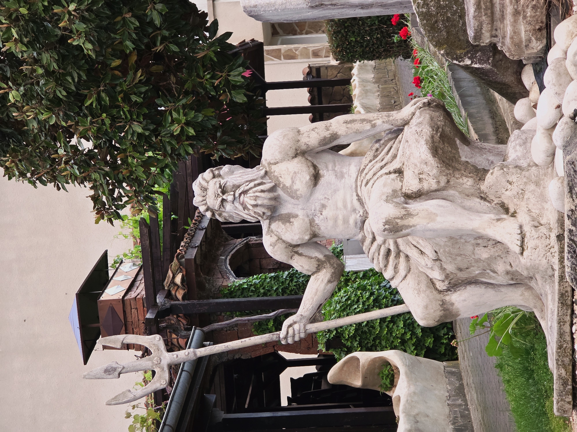 The image depicts a statue of a mythical male figure, possibly Neptune, holding a trident, surrounded by vegetation and architectural elements. The statue is made of light-colored stone, possibly marble or concrete, and shows signs of weathering or aging.
Behind the statue are various architectural elements, including a pergola covered with ivy and a brick structure. The scene appears to be set in an outdoor garden or courtyard, likely in a residential or commercial setting. There are also flowers and greenery in the background, adding to the garden-like ambiance.
The location is specifically in the city of Obzor, Bulgaria, based on the available information. The scene conveys a combination of classical art and natural elements in an outdoor environment, suggesting a garden or courtyard.