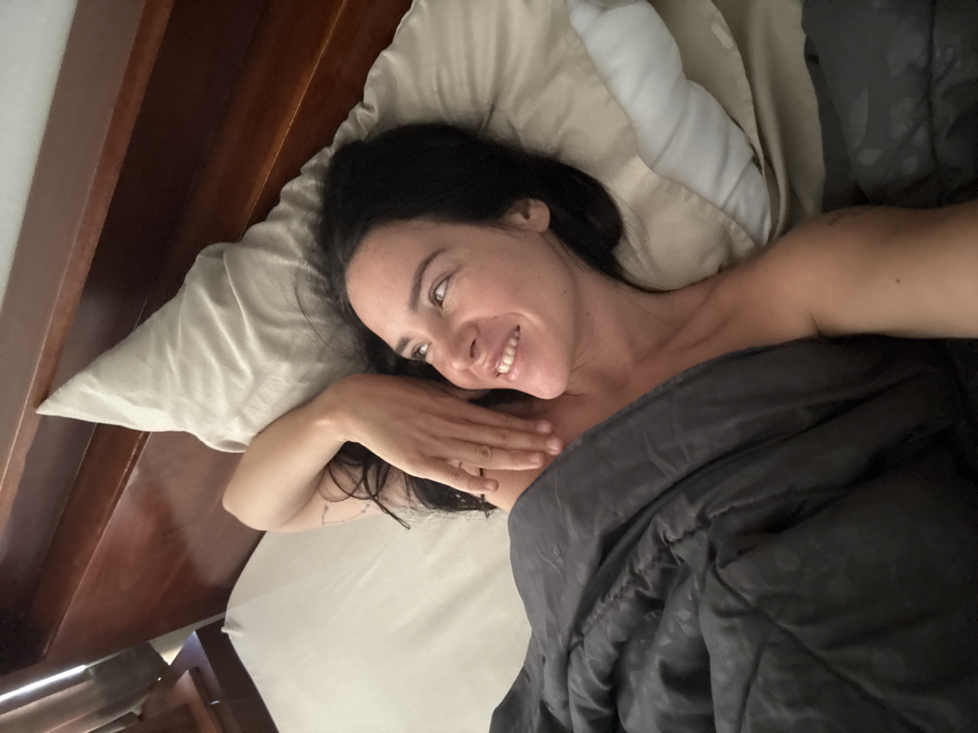 A woman with dark hair is lying in bed, partially covered by a dark grey comforter. She is propped up on a light-colored pillow, and her right arm is resting on her chest, with her hand near her face. Her head is turned to the left, and she is smiling and looking upwards. The bed appears to be in a room with a wooden headboard. The scene is consistent with a bedroom in North Kuta, Indonesia.