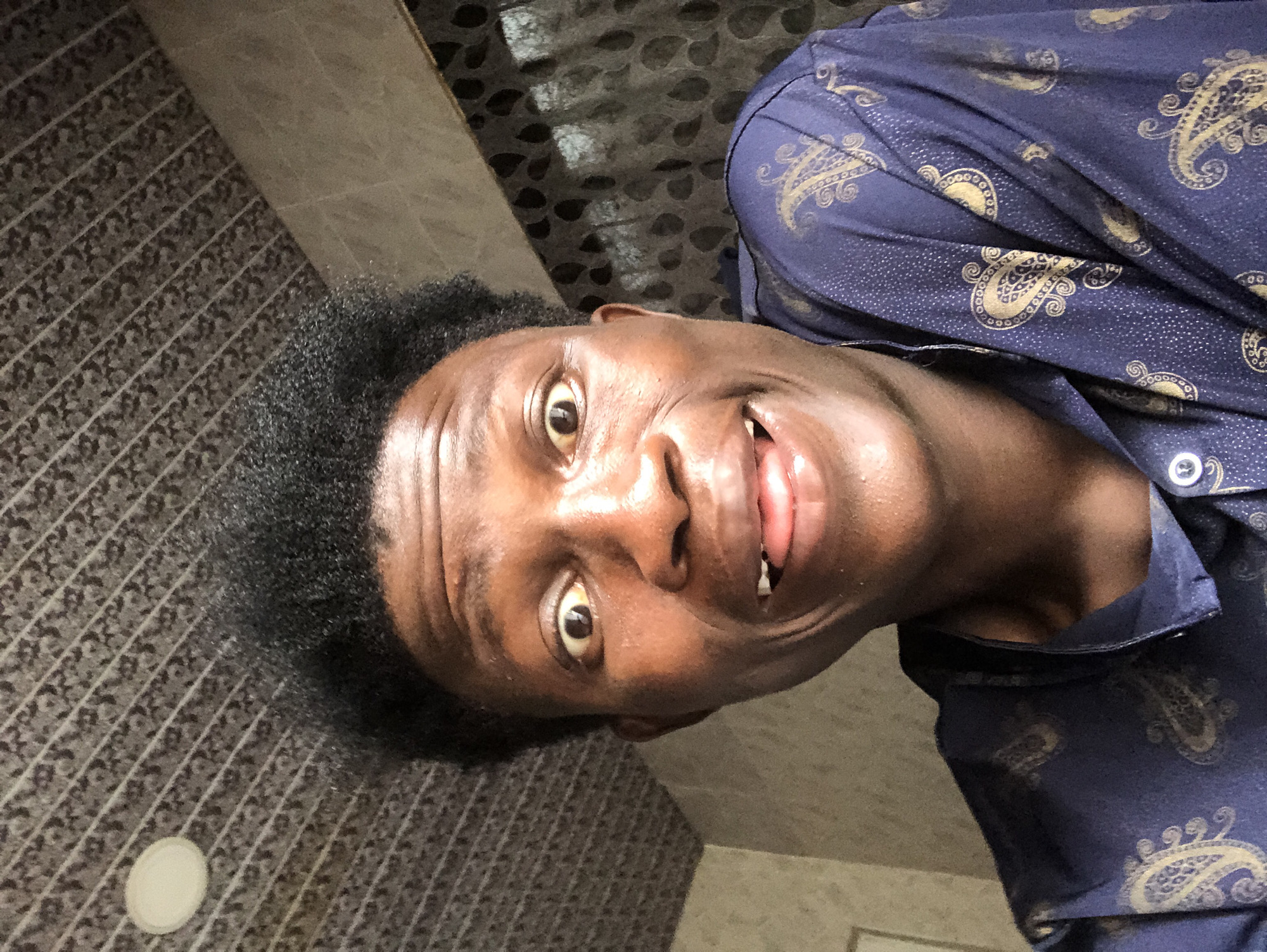 A close-up, eye-level, medium shot shows a young Black male in Ajah, Nigeria. He is facing the camera directly, his head tilted slightly back and to the left. His facial expression is one of surprise or excitement, with wide, startled eyes, raised eyebrows, and an open mouth with his tongue slightly visible. He has short, dark, curly hair and dark skin. He is wearing a dark blue shirt with a gold paisley pattern. The background consists of a patterned ceiling, a tiled wall, and a patterned curtain. The lighting appears to be natural, possibly from a window or overhead light. The image is a selfie, taken from a low angle.
