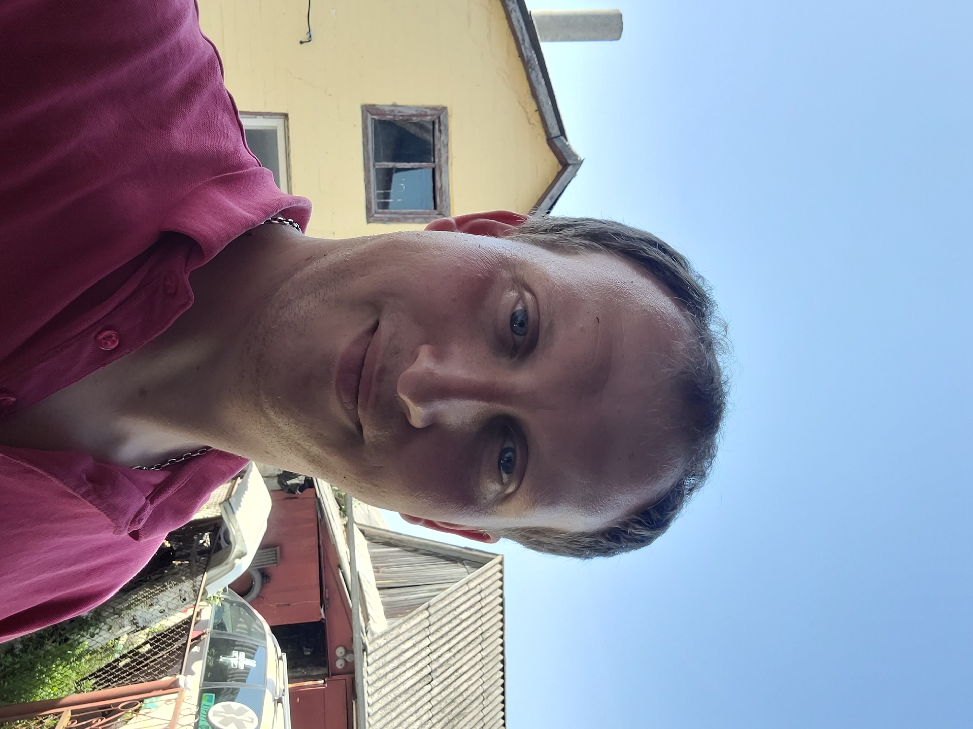 The media file is a selfie of a man with fair skin and light brown hair, taken outdoors during the daytime. The man is wearing a pink collared shirt and a silver chain necklace. He is looking towards the camera with a neutral expression.
The background is slightly out of focus, but appears to be a rural or industrial setting. Behind the man, a yellow building with a small window is visible. Further back, there is a glimpse of a roof with wooden shingles. In the lower portion of the image, parts of what might be vehicles or structures can be seen, along with some greenery and a metal fence. The sky is clear and blue, suggesting fair weather. The lighting indicates it is daytime, possibly late morning or early afternoon. No visible text is present in the image.