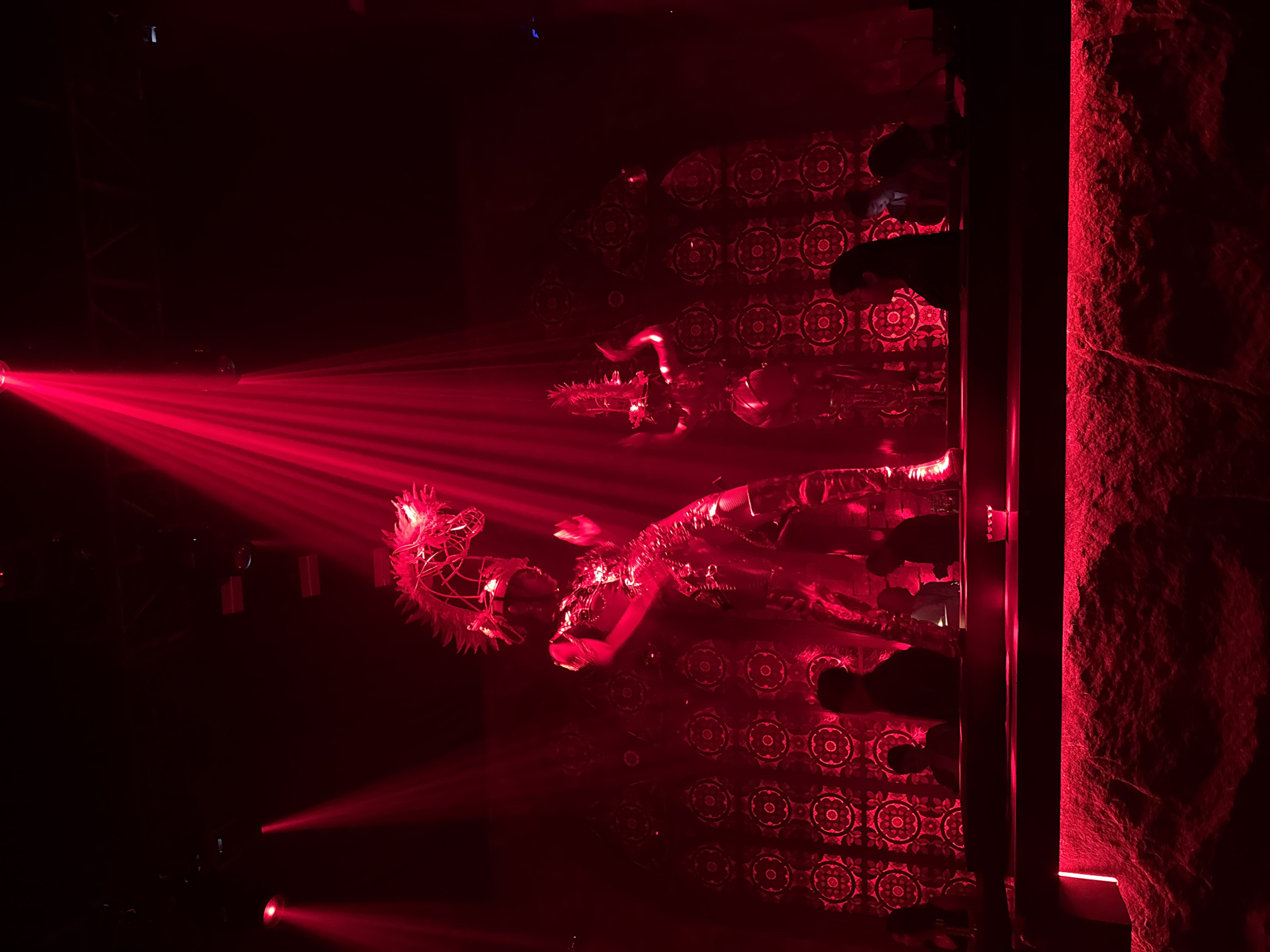 In a nightclub setting in North Kuta, Indonesia, the dimly lit space is illuminated by vibrant red lighting and theatrical spotlights. Two large, costumed performers stand prominently, bathed in the intense red beams. The performer on the left is taller, wearing an elaborate, metallic, insect-like costume with a large, spiky headdress resembling a dragon or mythical creature. The performer on the right is slightly shorter and also adorned in a metallic, costume, with what appears to be horns on their head. Behind them, a wall is decorated with a repetitive, circular, mandala-like pattern in a darker red. In the foreground, several patrons are visible, silhouetted against the illuminated backdrop, seated or standing at a bar. The bar itself features a textured, stone-like surface in the front, with red LED strip lighting along its base. The overall atmosphere is dramatic and visually striking, indicative of a nightlife entertainment venue.