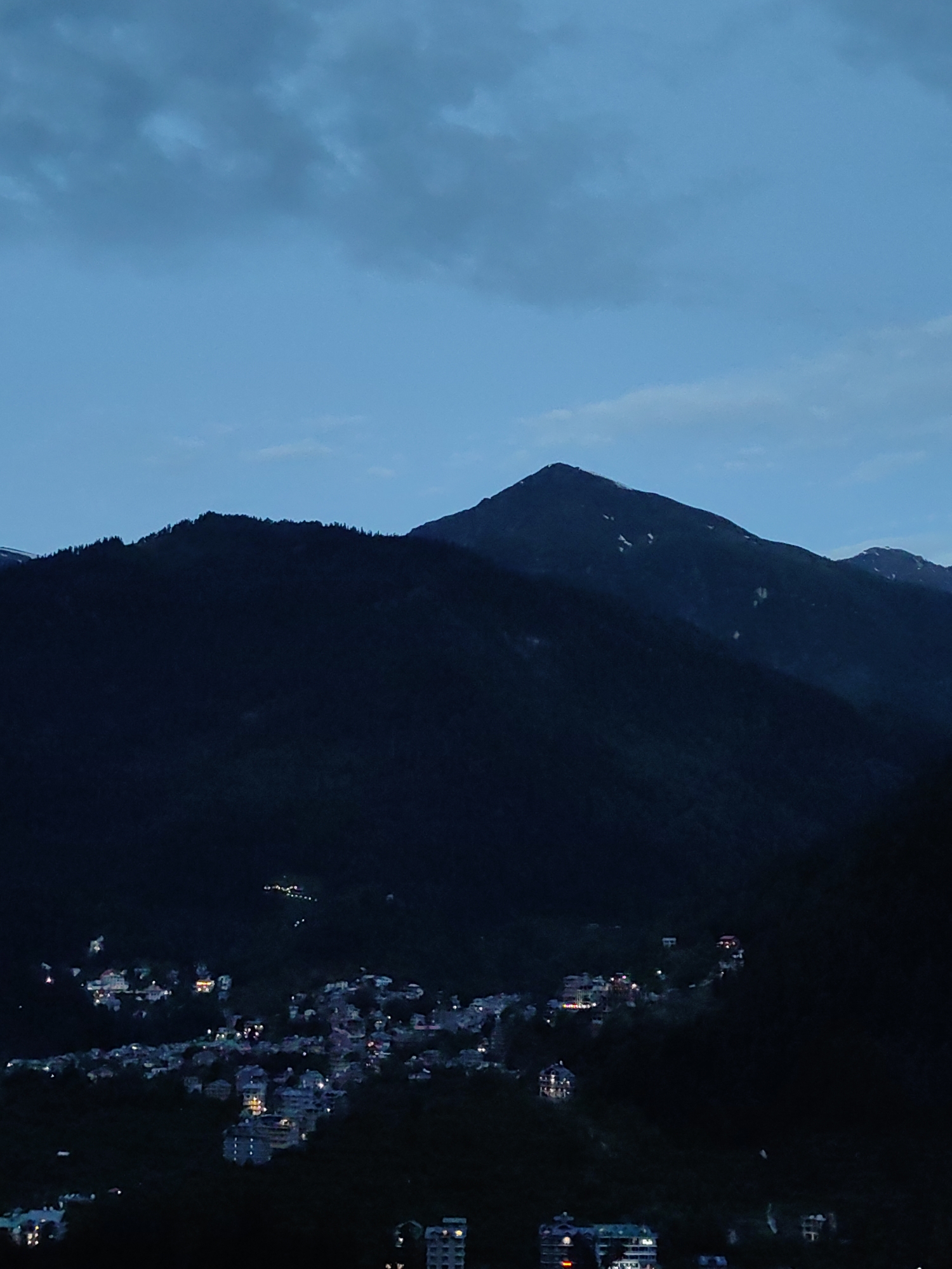 The image depicts a mountainous landscape at dusk, with a village nestled in the valley. The sky is a deep blue, transitioning to lighter shades near the horizon, with scattered clouds. The foreground is dominated by dark, silhouetted mountains covered in trees. In the mid-ground, a village is visible, illuminated by numerous lights from houses and buildings. The village appears to be spread across the slopes of the mountain. The overall impression is one of a tranquil, remote mountain settlement as night begins to fall. This scene is consistent with the location of Vashisht, India, a village known for its mountainous terrain and hot springs.