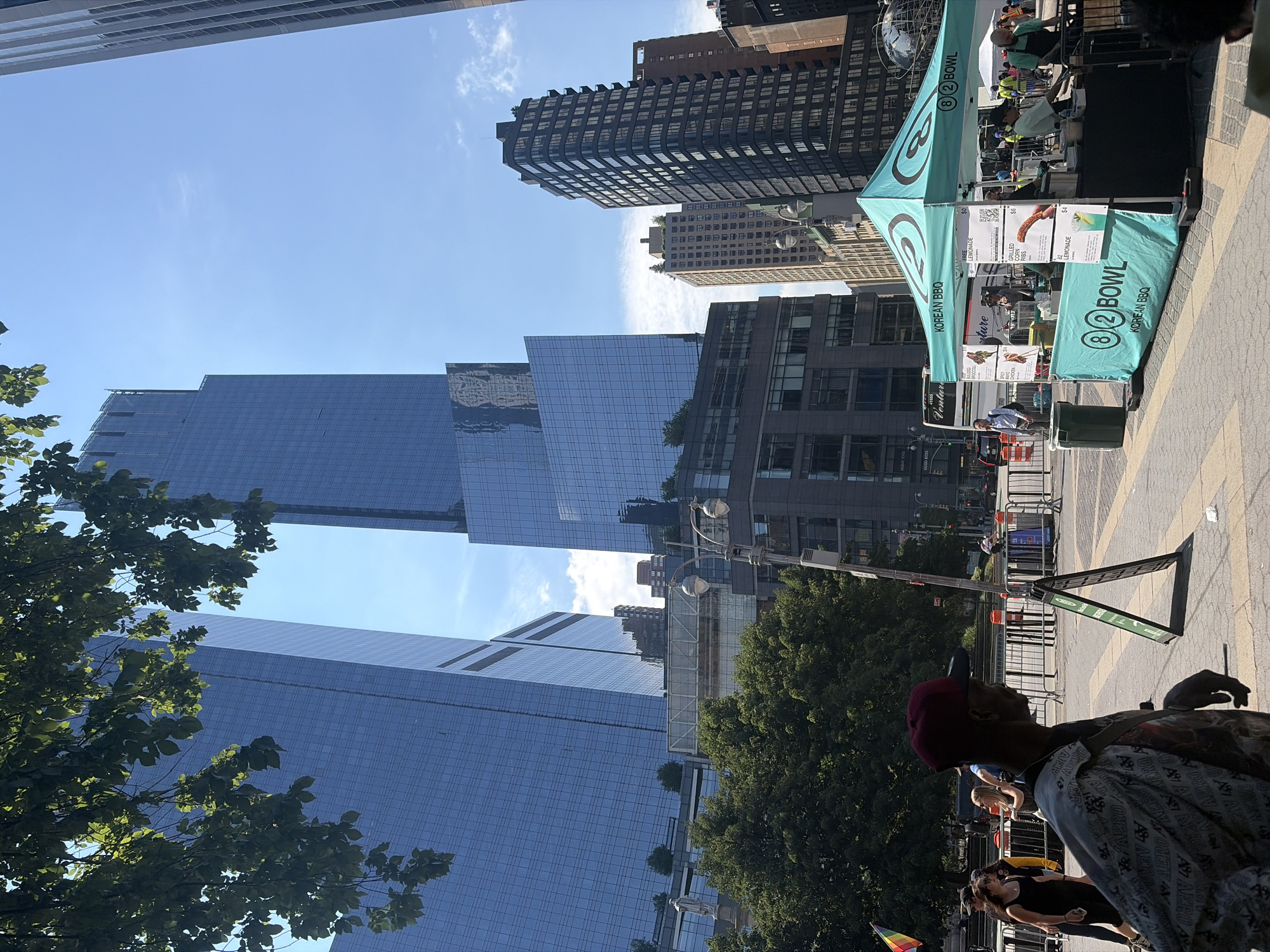 In New York City, on a sunny day, a street scene unfolds with a focus on a food stall. The stall, a turquoise tent with "8 2 BOWL KOREAN BBQ" branding, is set up on a paved plaza. Various food items, including grilled corn, spicy bulgogi chicken, and lemonade, are advertised on the menu boards. A person in a light green shirt and a person in a black apron are working behind the counter. In the foreground, a person wearing a red cap and a patterned jacket is walking away from the camera. Several other individuals are visible in the background, some walking and others standing near a rainbow flag. The scene is bordered by tall glass-fronted buildings, suggesting an urban environment. The sky is clear blue with a few scattered clouds.