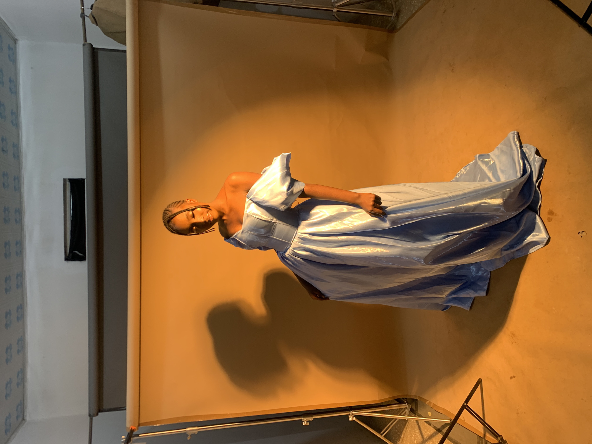 A young Black woman with braided hair stands center frame, looking down and to her right, in front of an orange backdrop. She is wearing a light blue, off-the-shoulder gown with puffy sleeves and a full skirt. The lighting creates a dramatic shadow of the woman on the backdrop to her left. In the foreground, a light stand is visible on the left, and another on the right. The ceiling above the backdrop appears to be a drop ceiling with a patterned tile design. The location is identified as Gura Top, Nigeria.