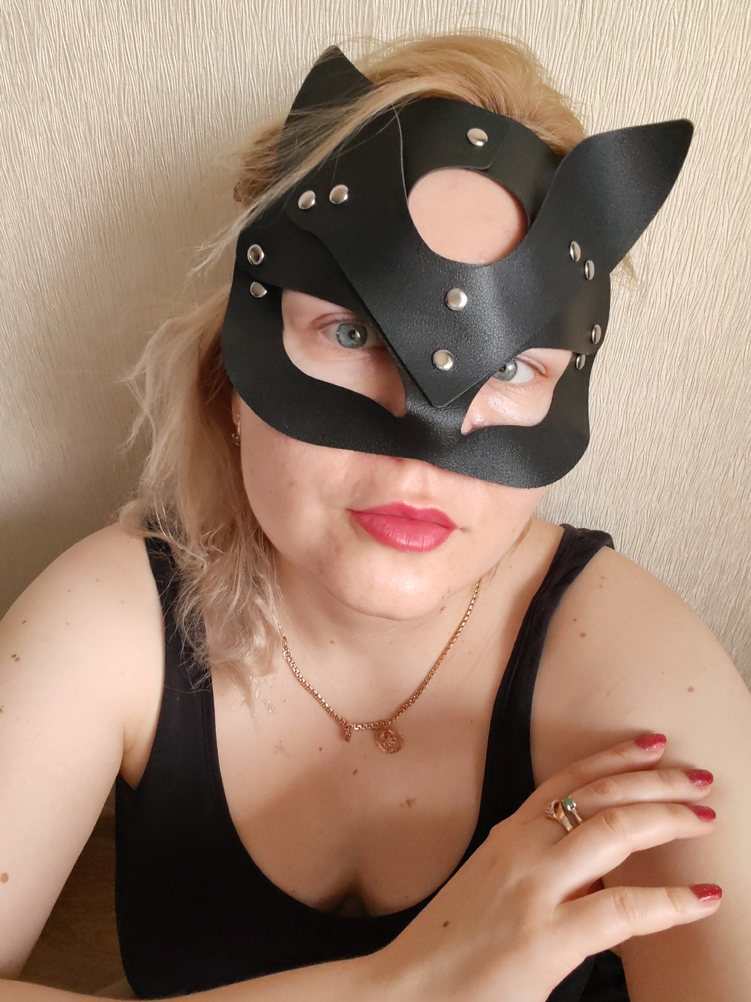 A woman with blonde hair and fair skin is wearing a black leather cat mask with metal studs. The mask covers the upper half of her face, leaving her eyes and mouth visible. She has light blue eyes and wears red lipstick. She is wearing a black tank top and a gold necklace with a circular pendant. She has a gold ring with a green stone on her right hand, and her nails are painted red. She is positioned in front of a neutral-colored wall. The image does not provide sufficient information to ascertain a specific location within Izhevsk, Russia.