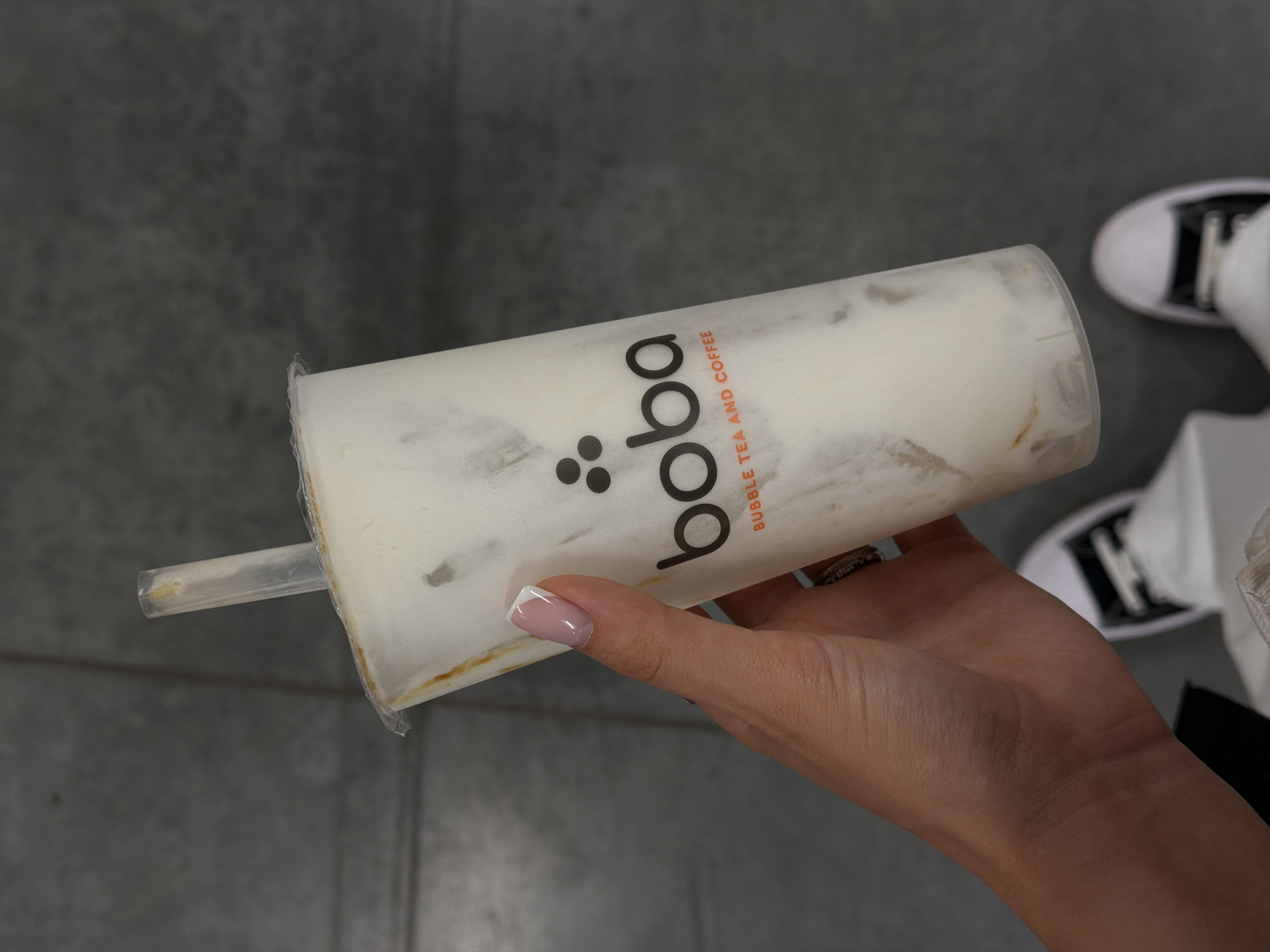The image shows a hand holding a plastic cup of boba tea. The cup has the word "boba" printed on it, along with "bubble tea and coffee". A clear plastic straw is inserted into the cup. The hand has a ring on the ring finger and a manicure with white tips. In the background, parts of two shoes (white with black stripes) and clothing are visible. The floor appears to be gray concrete. There is no indication of the city, Kazan, Russia.