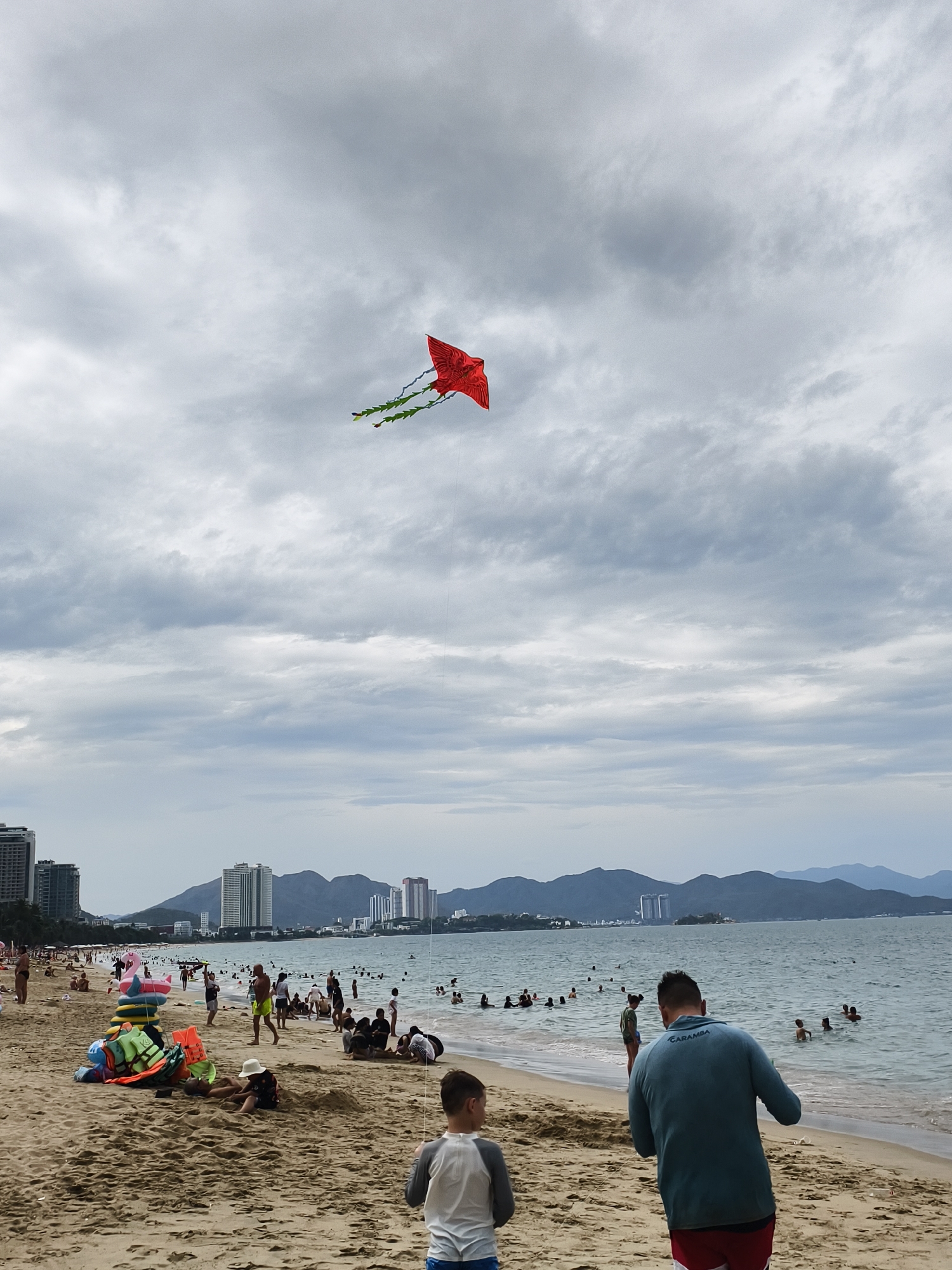 This media file depicts a busy beach scene under an overcast sky. A striking red kite, shaped somewhat like a bird or dragon and adorned with green streamers, is prominently flying high in the cloudy sky.
The sandy beach is bustling with numerous people engaging in various leisure activities. Many individuals are seen relaxing, sunbathing, or playing in the sand. In the foreground, a child wearing a white hat is seated on the sand, possibly digging, near a stack of colorful life vests and a large inflatable pink flamingo. Several people are also enjoying the ocean, wading or swimming in the calm, greyish-blue water.
Two figures are visible in the immediate foreground, with their backs to the viewer. A man in a teal long-sleeved shirt has the word "CARAMBA" printed on its back, and a young boy in a white and grey long-sleesleeved top stands beside him. They appear to be looking towards the ocean, and the kite string is faintly visible extending from the sky towards their general direction.
In the background, a coastal city is visible, characterized by several high-rise buildings lining the shore. Beyond the city, a range of hazy mountains stretches across the horizon. The sky is predominantly covered with thick, grey clouds, suggesting an overcast day, but the beach is still full of activity. The scene conveys a popular holiday or recreational spot.