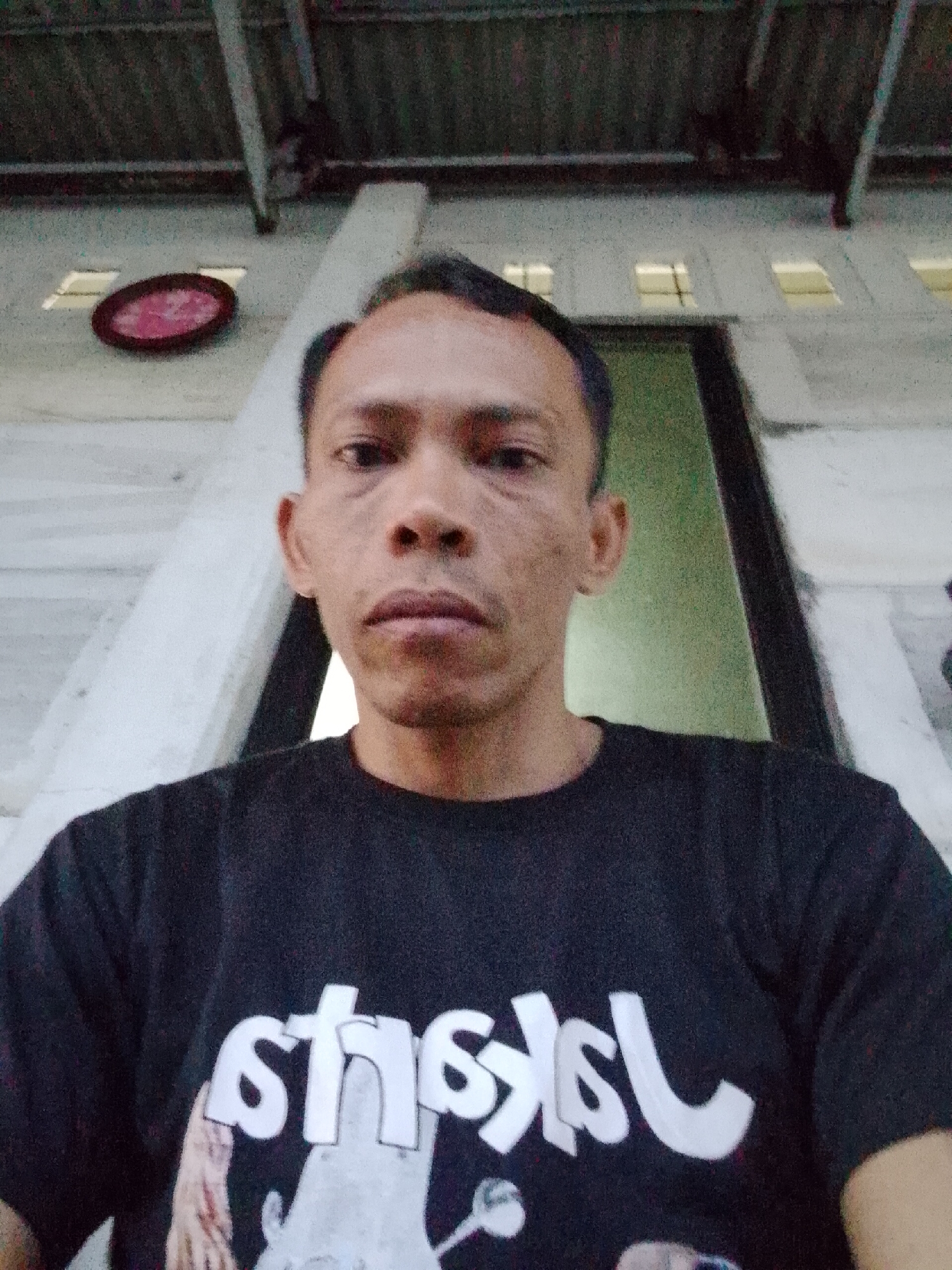 The image is a selfie of a man. He is wearing a black t-shirt with white lettering and a partial image on it. He appears to be standing on a porch or covered area of a building. The background includes a white wall with a few small windows near the top, a dark green doorway, a red circular object on the wall, and a gray ceiling. The setting appears to be in Bukit Ketok, Indonesia based on the camera’s location. No specific actions or interactions are observable in the image.
