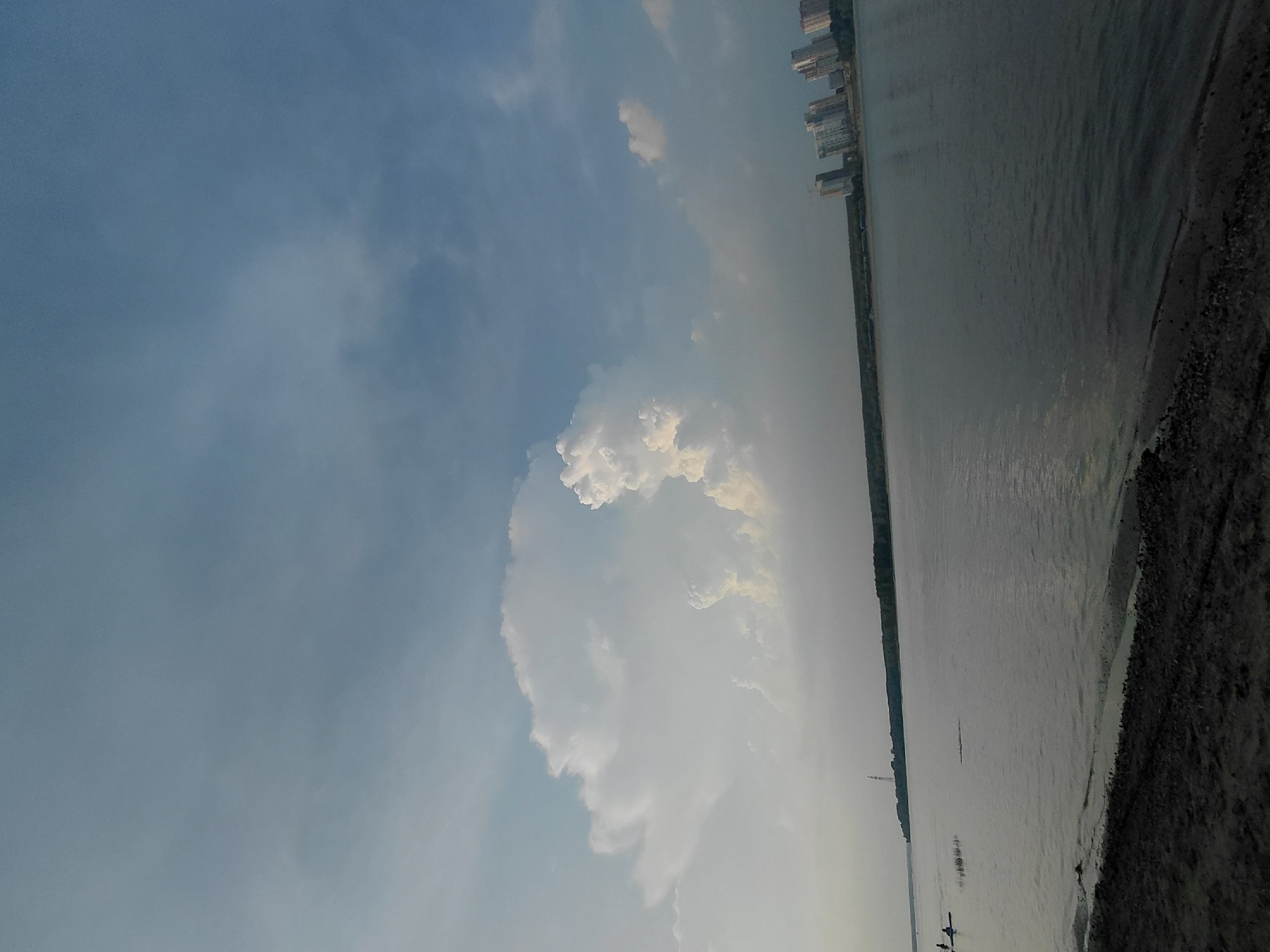 The image depicts a wide river, likely the Ob River, on a cloudy day in Novosibirsk, Russia. The foreground shows a sandy shore with dark pebbles. Across the river, a treeline separates the water from a cluster of tall, modern buildings, possibly apartment complexes, on the horizon. To the left, two individuals are visible in the water, one appearing to be in a kayak. A distant tower is also visible along the shoreline. The sky is filled with dramatic, white cumulus clouds against a pale blue background.