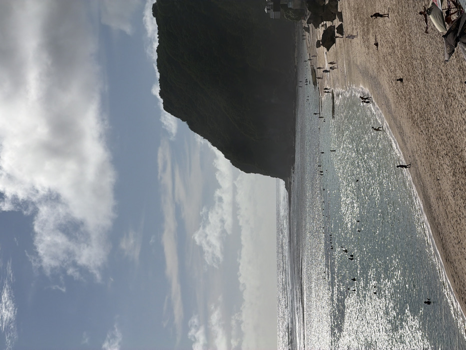 This image depicts Melasti Beach in Ungasan, Indonesia. The scene presents a wide expanse of light beige sandy beach in the foreground, populated by approximately 25 individuals engaged in walking, standing, or relaxing. One person lies supine in the sand in the bottom right corner. Adjacent to the beach is the ocean, shimmering with reflected sunlight, where an estimated 30 individuals are observed swimming or wading. A submerged reef or rock formation is visible between the shallow water and more distant breaking waves. The background on the right is dominated by a large, steep, dark green cliff heavily covered in vegetation, which meets the coastline directly. Several small, light-colored structures are built into the lower portion of this cliff face. The sky above is light blue with scattered white and grey cumulus clouds. In the bottom right foreground, partially visible beach umbrellas and a watercraft are present.