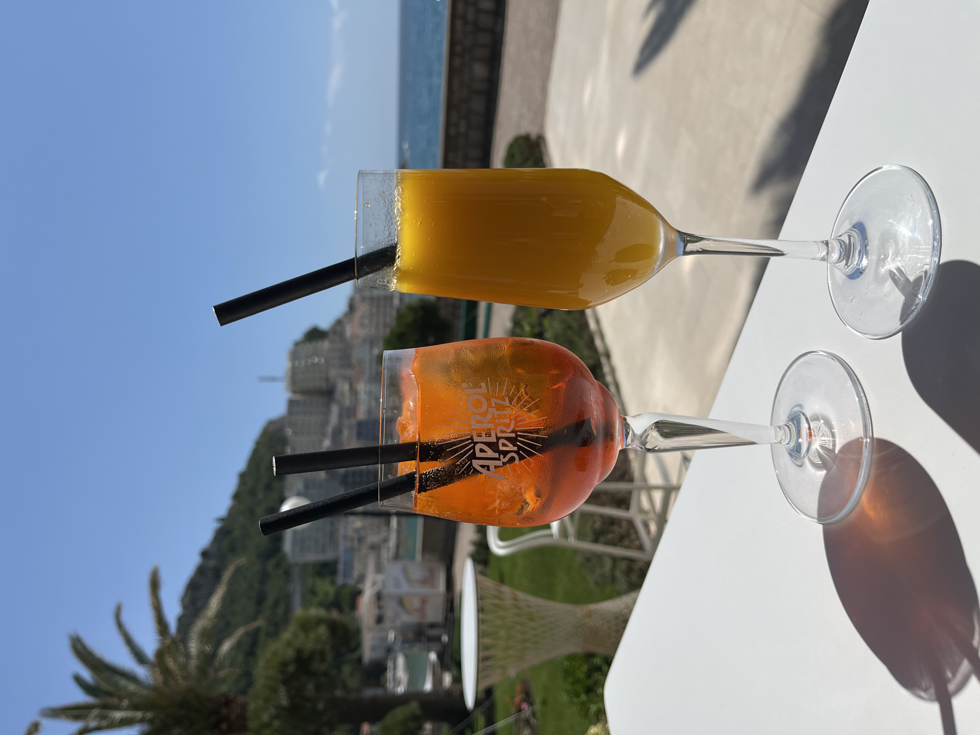 Two cocktails are on a white table against a blurred backdrop of the city of Bečići, Montenegro, the sea, and a clear blue sky. The lower cocktail is an Aperol Spritz with two black straws and the "Aperol Spritz" logo on the glass. The top cocktail is a yellowish-orange color, also with a single black straw. Both cocktails are in tall glasses with a stem and a round base. The table casts shadows.