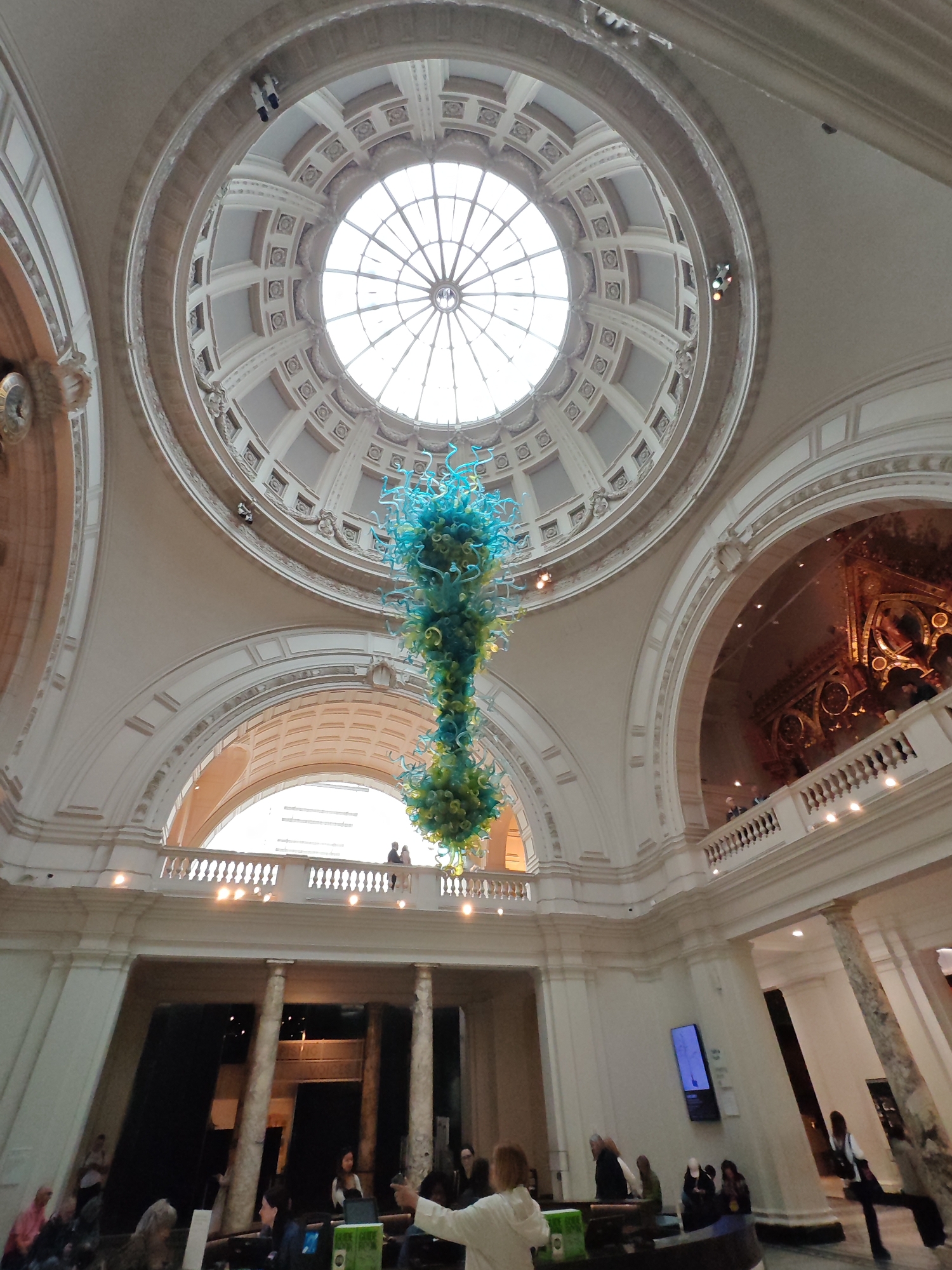The image depicts the interior of the Victoria and Albert Museum in London, United Kingdom. The scene is dominated by the grand hall's architectural features: a large dome with a central skylight, arched openings leading to other galleries, and ornate detailing on the walls and ceilings. A large, colorful glass sculpture hangs from the center of the dome. In the foreground, a group of people are gathered, some seated and some standing. A woman in a white jacket appears to be gesturing or speaking to the group. Beyond the group, there are columns and dark openings, possibly leading to exhibition spaces. On the upper level, along a balcony, several people are visible. The overall impression is one of a spacious, ornate museum hall with visitors engaged in activities.
