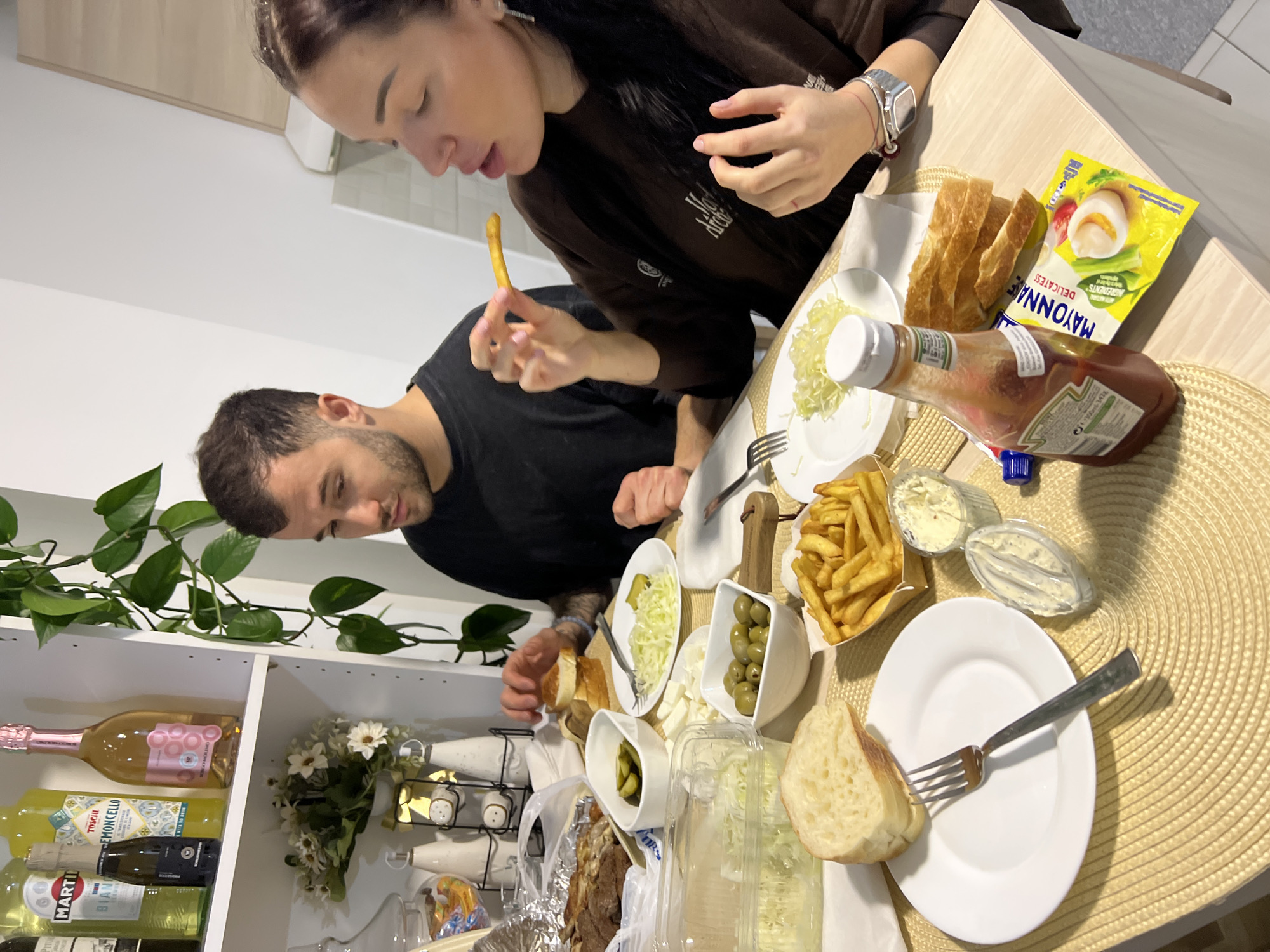 Two individuals, a male and a female, are seated at a table indoors, engaged in eating a meal. The table is set with various food items including bread, fries, olives, salad, and condiments such as ketchup and mayonnaise. The male, wearing a black shirt, is looking down at the food. The female, wearing a brown sweatshirt and a watch, is holding a french fry and appears to be about to eat it.
In the background, there is a white shelving unit displaying bottles of alcohol and decorative items. A potted plant sits on the left side of the frame.
The image does not contain enough information to confirm its location as Boreti, Montenegro.