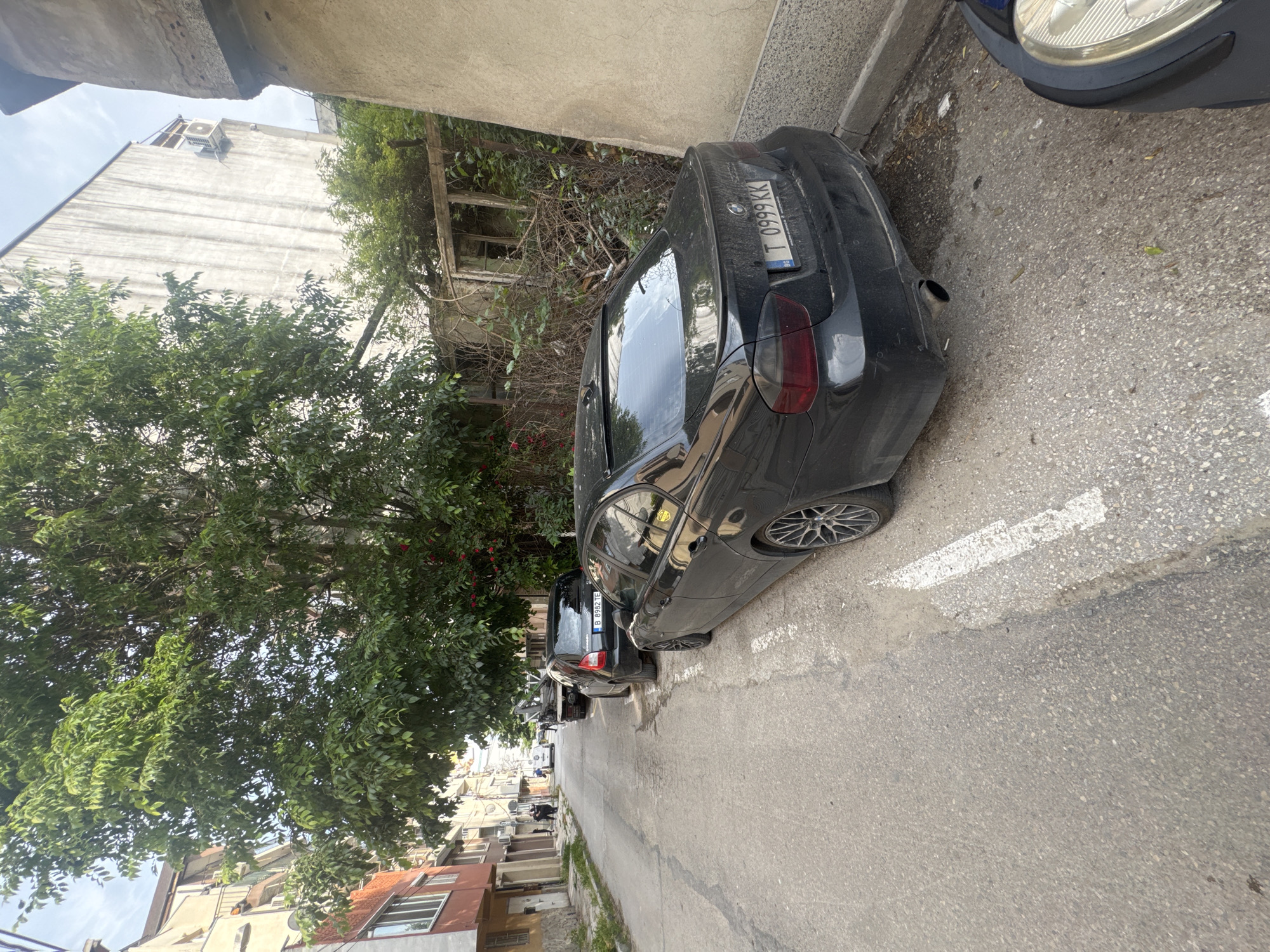 This daytime image depicts a narrow street in an urban area, likely Varna, Bulgaria, characterized by parked cars, varied architecture, and lush greenery. The perspective looks down the street, with buildings on both sides.
In the foreground, a dusty black BMW sedan is prominently parked parallel to the curb. Its rear is visible, showing a license plate with the registration "T 0999 KK". The car features aftermarket multi-spoke wheels and a single exhaust pipe. Immediately in front of this vehicle, only the blue bumper and part of a headlight of another car are visible. Behind the black BMW, another dark-colored sedan is parked, displaying the Bulgarian license plate "B 8982 TE".
To the right of the parked vehicles, a severely dilapidated building with broken windows and dense, overgrown vegetation is visible, suggesting abandonment. Further along the street, various multi-story buildings with different facades line the road, some appearing residential. One building features an external air conditioning unit. Large, leafy trees overhang the street, providing shade, with some displaying small red berries or flowers.
The asphalt street surface is worn, showing cracks and patches. A faded white solid line road marking is visible on the pavement near the parked cars. The sky above is partly visible, appearing hazy blue or lightly cloudy. No specific human activity is clearly ongoing, though a distant, blurry figure can be seen further down the street. The Bulgarian license plates confirm the geographical location.