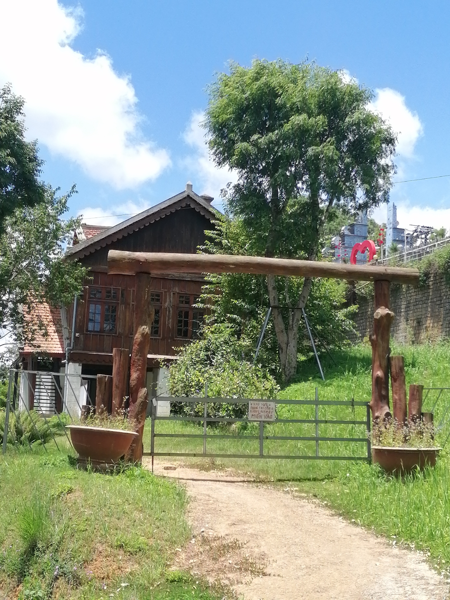 The image depicts the entrance to a property, likely a residence or a tourist attraction, situated in Da Lat, Vietnam. A rustic wooden archway, constructed from a thick log supported by two upright wooden posts, spans a dirt pathway. This archway frames a metal gate, partially open, which leads to the property. On either side of the gate, large, shallow terracotta pots serve as planters, with one on the left containing sparse greenery and the other on the right adorned with small flowering plants.
Beyond the gate, a traditional wooden house with a pitched, tile roof is visible. The house features multiple windows with dark frames. The environment surrounding the property is lush and green, with abundant vegetation including trees and grass. In the background, a stone wall can be seen, and further in the distance, elements of a city skyline are present, including buildings and what appears to be a decorative red heart sculpture. The sky is blue with scattered white clouds, indicating a clear day. The overall impression is one of a serene, possibly historical, location in Da Lat.