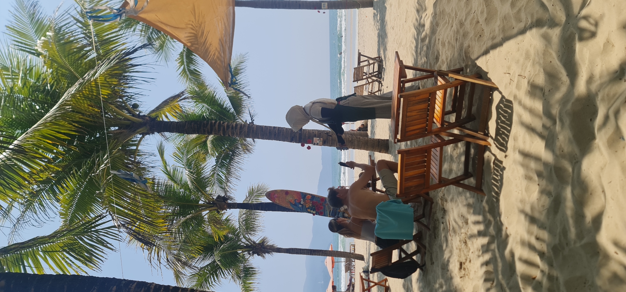 The image depicts a beach scene in Đà Nẵng, Vietnam. Palm trees frame the left side of the image, while a sandy beach dominates the right. In the background, the ocean meets the horizon under a clear sky.
Several people are present. A woman wearing a head covering and apron appears to be working, possibly serving customers. A man is seated in a wooden chair, looking at a phone. Another person is seated next to him.
Wooden chairs are arranged on the sand, suggesting a beachside establishment. A surfboard decorated with flowers leans against a palm tree. A sunshade is partially visible above.