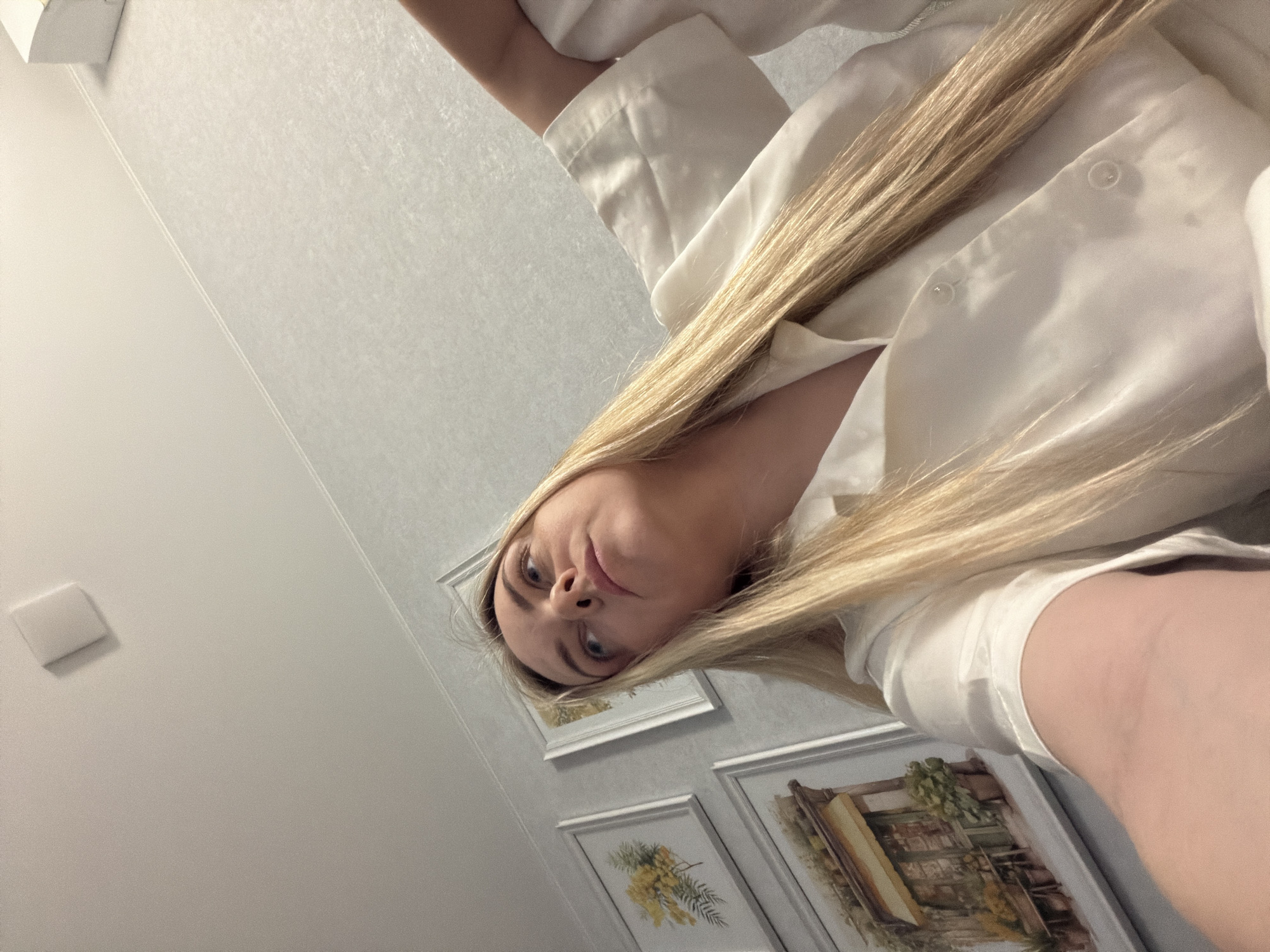 A selfie-style photograph captures a young woman from an inverted, low-angle perspective, making her appear upside down. She has long, straight, blonde hair that cascades downwards in the frame, and fair skin. Her blue eyes are open, looking directly towards the camera, with a neutral to slightly pensive expression, featuring a subtle pout. She is wearing a light-colored, possibly white or off-white, collared button-up shirt, visible around her neck, shoulders, and upper torso, with some buttons clearly visible.
The background consists of a light-colored wall, likely wallpapered, featuring a subtle textured pattern. On the wall behind and below her head (from the viewer's perspective), there are several framed pictures. One prominent frame contains artwork depicting yellow flowers, possibly mimosa. Another frame, partially visible, shows a rustic or architectural scene, potentially with grapes or vines. A small, square, light-colored object, possibly a light fixture, vent, or sensor, is mounted on a plain light-colored surface in the upper left of the image. The lighting appears soft and even, suggesting an indoor setting. The overall scene conveys an intimate, casual moment. The location is Votkinsk, Russia. There is no visible text in the image.