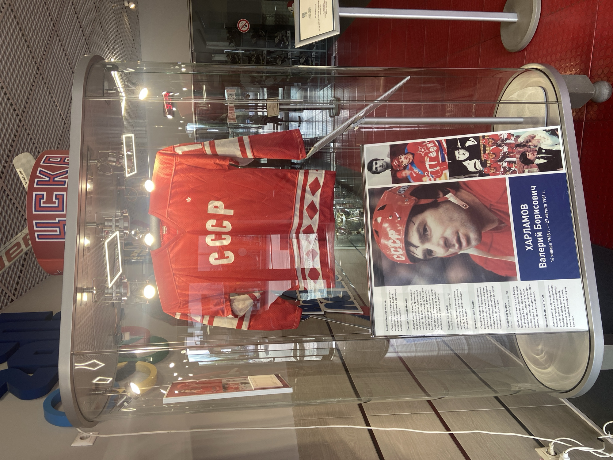 The image shows a display case containing a Soviet Union hockey jersey, a hockey stick, and a poster of Valeri Kharlamov, a famous Soviet hockey player. The poster includes Kharlamov's name and dates (January 14, 1948 – August 27, 1981), as well as quotes about him from other hockey figures. The jersey is red with white stripes and the Cyrillic letters "CCCP." The display case also features the letters "ЦСКА" in large, red lettering. The location is likely a museum exhibit about Soviet hockey or Valeri Kharlamov, possibly in Peresvet, Russia.