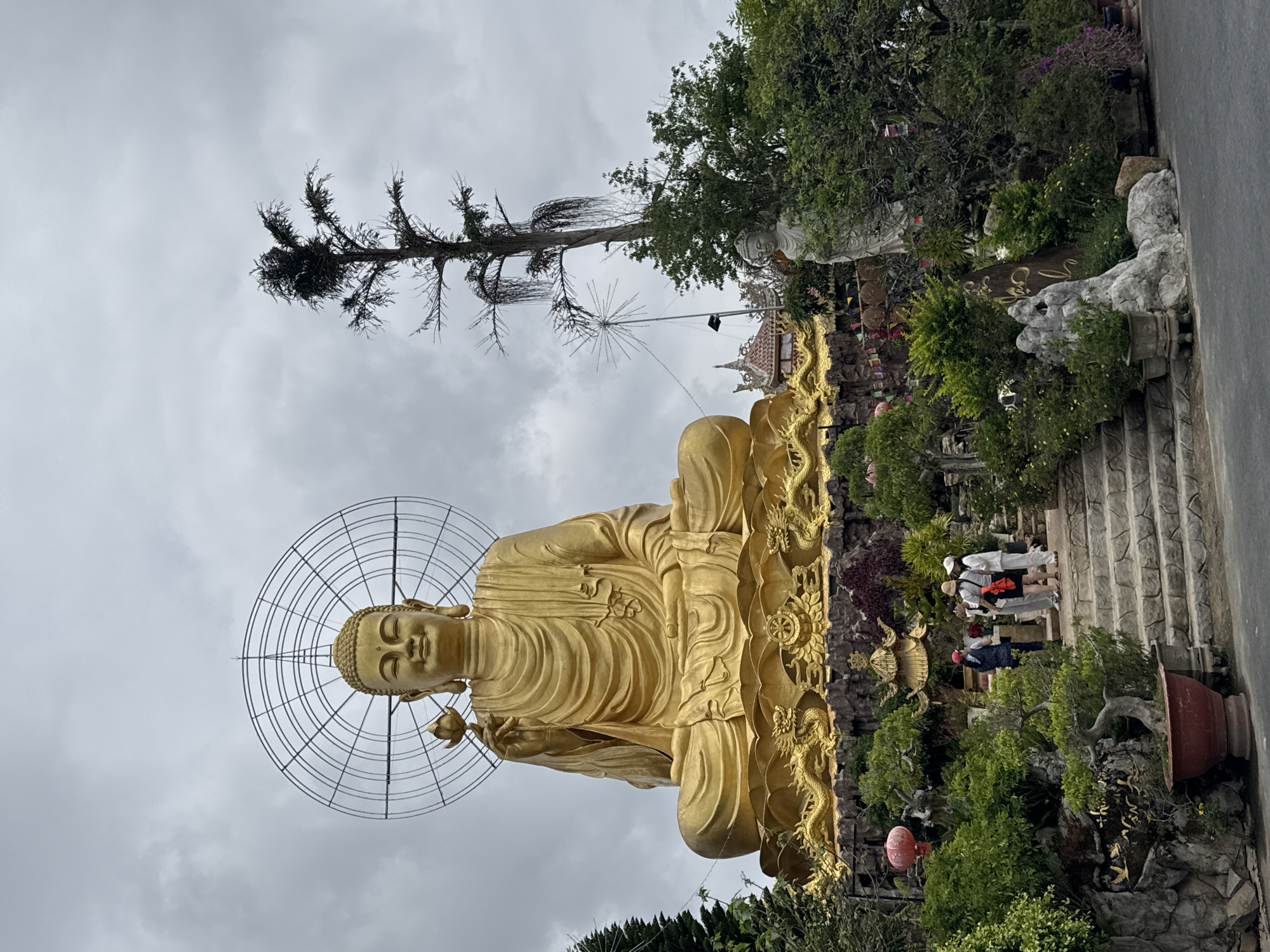 A large, majestic golden Buddha statue sits prominently outdoors at a temple complex in Lâm Viên, Đà Lạt, Vietnam, under an overcast sky. The Buddha is depicted in a meditative pose, holding a lotus flower in its right hand, with a large, elaborate metal halo or aureola behind its head. The statue rests on an ornate golden base adorned with intricate dragon sculptures and Buddhist motifs like the Dharma wheel and lotus blossoms.
To the right of the statue, a set of stone stairs ascends a lush, terraced garden. Several people, seemingly visitors or devotees, are seen walking on these stairs, observing the surroundings. The garden features diverse greenery, including potted bonsai trees, bushes, and larger trees, some adorned with small flags or banners. In the upper part of the scene, behind the Buddha, parts of traditional temple architecture with ornate roofs are visible. A tall, somewhat sparse tree stands out against the cloudy sky in the upper left background. Various stone formations, including one with unidentifiable golden script, are integrated into the landscaping along the stairs. A red lantern is visible near the base of the stairs. The weather appears cloudy, indicating either a mild or overcast day without direct sunlight.