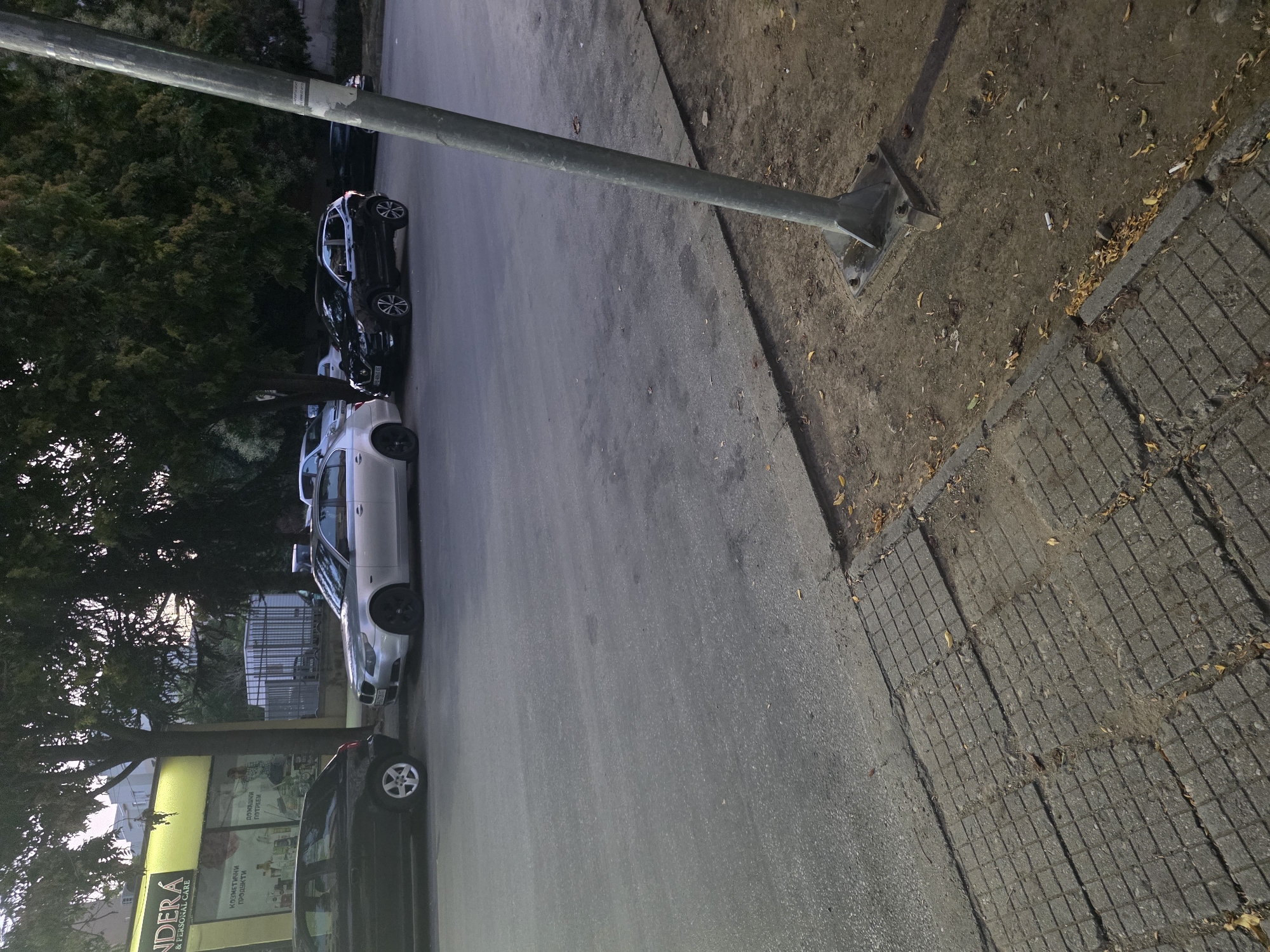 In Plovdiv, Bulgaria, a city scene unfolds with parked cars and urban infrastructure. The perspective is from street level, looking across a paved road.
In the foreground, a damaged sidewalk is visible, composed of square concrete slabs with visible cracks and missing sections. Dry leaves are scattered across the sidewalk and the adjacent dirt area. A metal pole, likely a street lamp or utility pole, stands prominently on the right side, its base bolted to a metal plate secured to the ground.
Further back, several cars are parked along the curb. On the left, a dark-colored hatchback is parked. To its right, a silver sedan is parked, followed by a black SUV. In the background, trees with dense foliage provide shade, and a building with a yellow facade, possibly a storefront, is visible. A metal fence can also be seen behind the cars. The lighting suggests it is either early morning or late afternoon, with soft shadows indicating diffused sunlight. The environment appears to be an ordinary street in a residential or commercial area of the city.