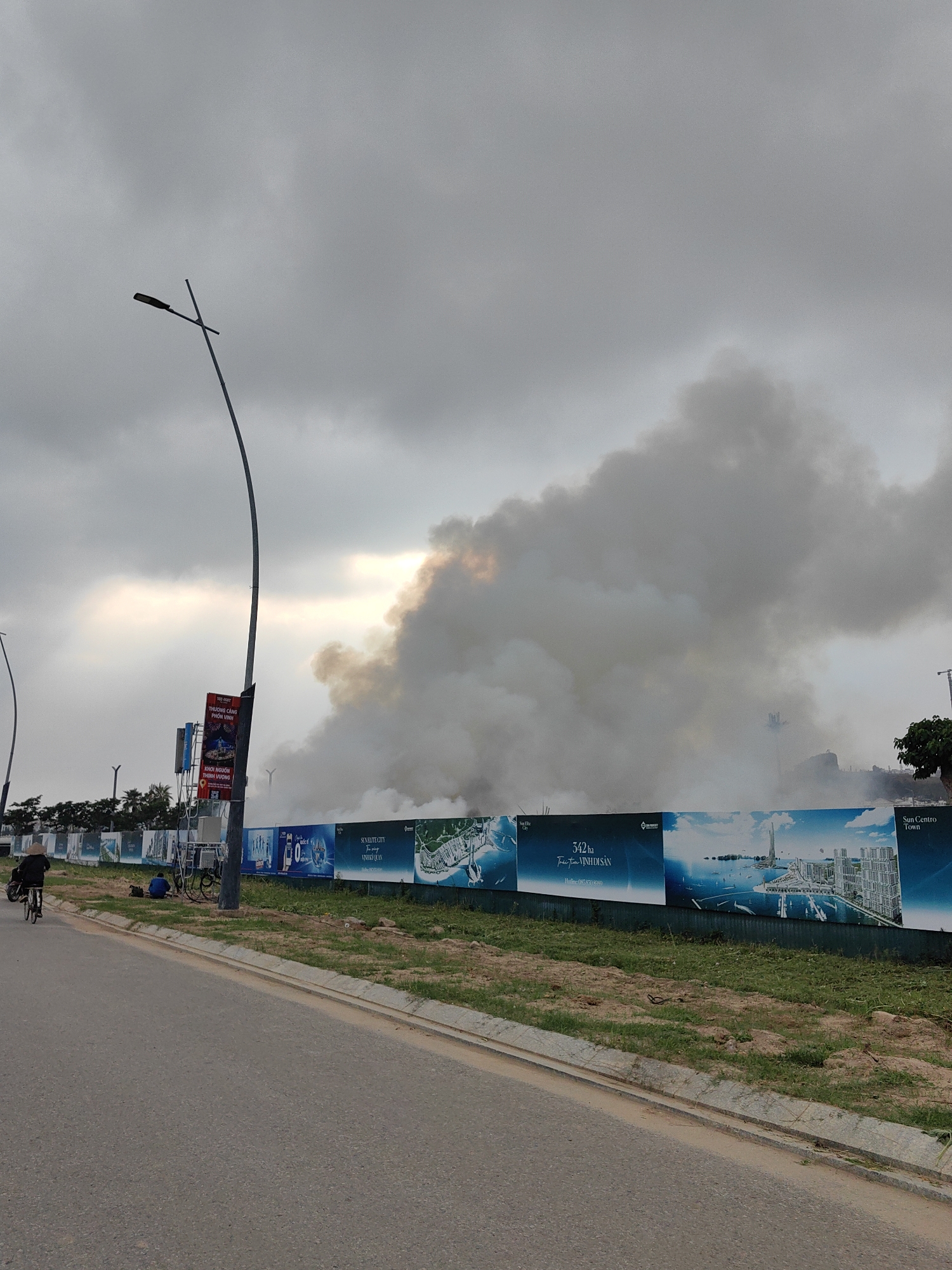 The image depicts a street scene in Bai Chay Ward, Vietnam. A plume of smoke rises into a cloudy sky behind a construction site. The site is bordered by a fence covered with promotional posters for "SunElite City" and "Sun Centro Town," advertising "342 ha Trai Tim Vinh Di San." A lamppost stands to the left of the smoke. A person on a bicycle rides along the asphalt road that borders the construction site. Another individual sits on the grass near the fence.