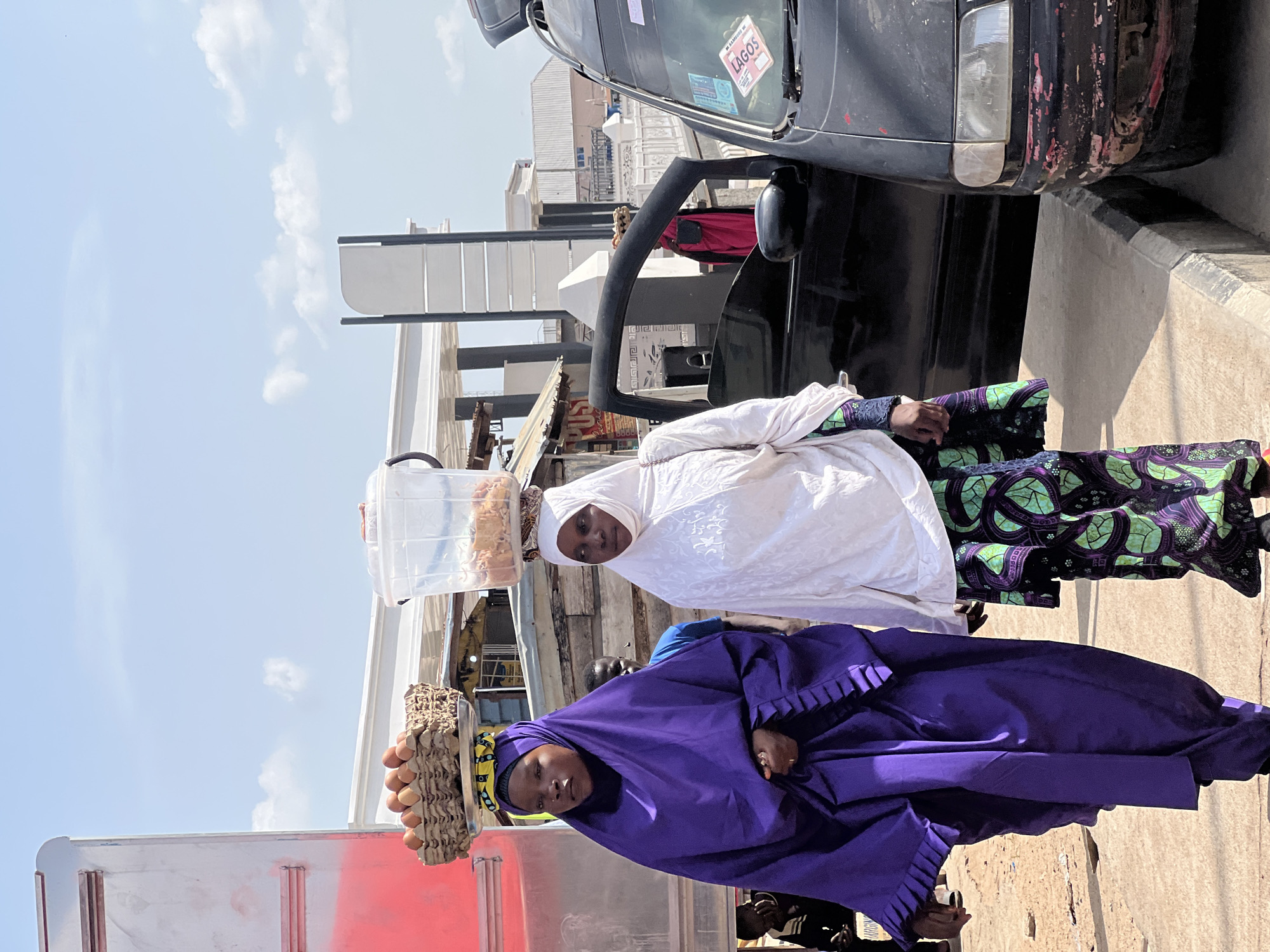 Two women are walking down a street in Jalingo, Nigeria. The woman on the left is wearing a purple abaya and a hijab. She is carrying a basket of eggs on her head. The woman on the right is wearing a white hijab and a patterned skirt. She is carrying a clear plastic container filled with food items on her head. A black van is parked to the right of the women. The street is paved and there is a sidewalk. The sky is blue with some clouds. The scene appears to be a busy market or street.