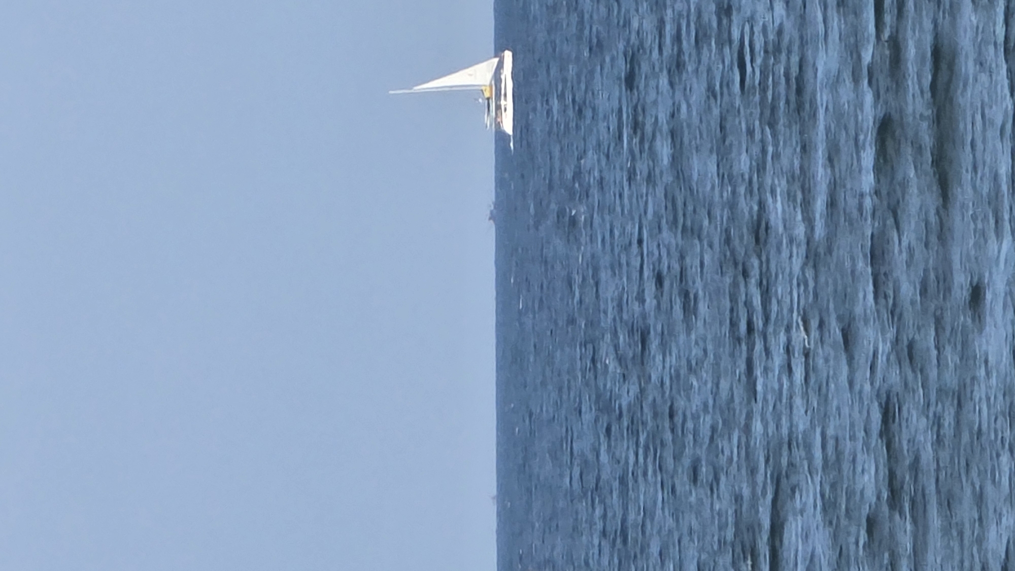 The image shows a sailboat on the water, with a light blue sky in the background. The water appears textured, with vertical lines indicating ripples or waves. The sailboat is white with a triangular sail. The image orientation is rotated. The location is not specified, nor is the city of Nha Trang, Vietnam.