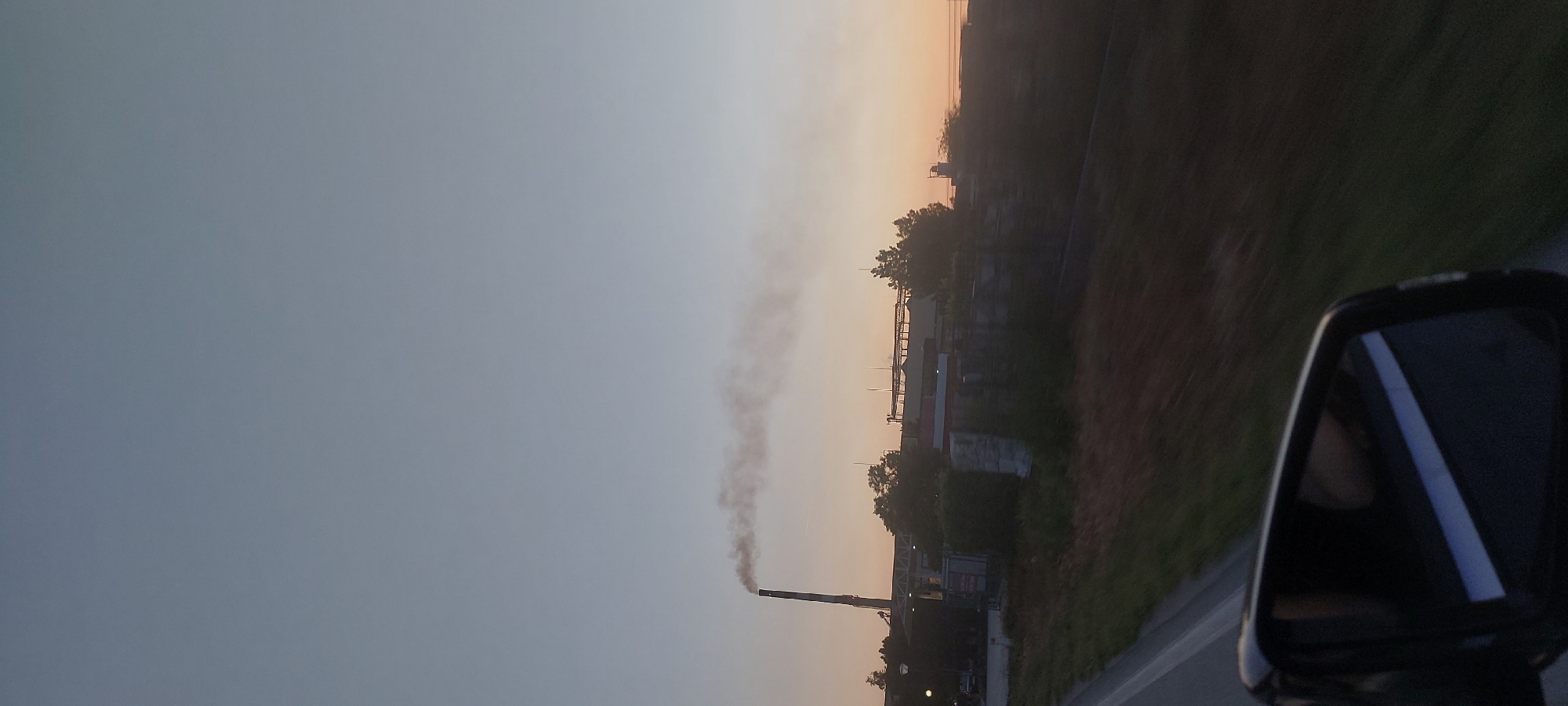 A photograph shows a factory spewing smoke from a tall smokestack near Katunitsa, Bulgaria. The image was likely taken from a moving vehicle, as evidenced by the blurred motion and the inclusion of a side-view mirror in the frame. The sky is a gradient of colors, suggesting either sunrise or sunset. A road is partially visible in the lower right, along with some vegetation. The factory appears to be an industrial complex with various structures and piping. The overall scene suggests industrial activity impacting the environment.