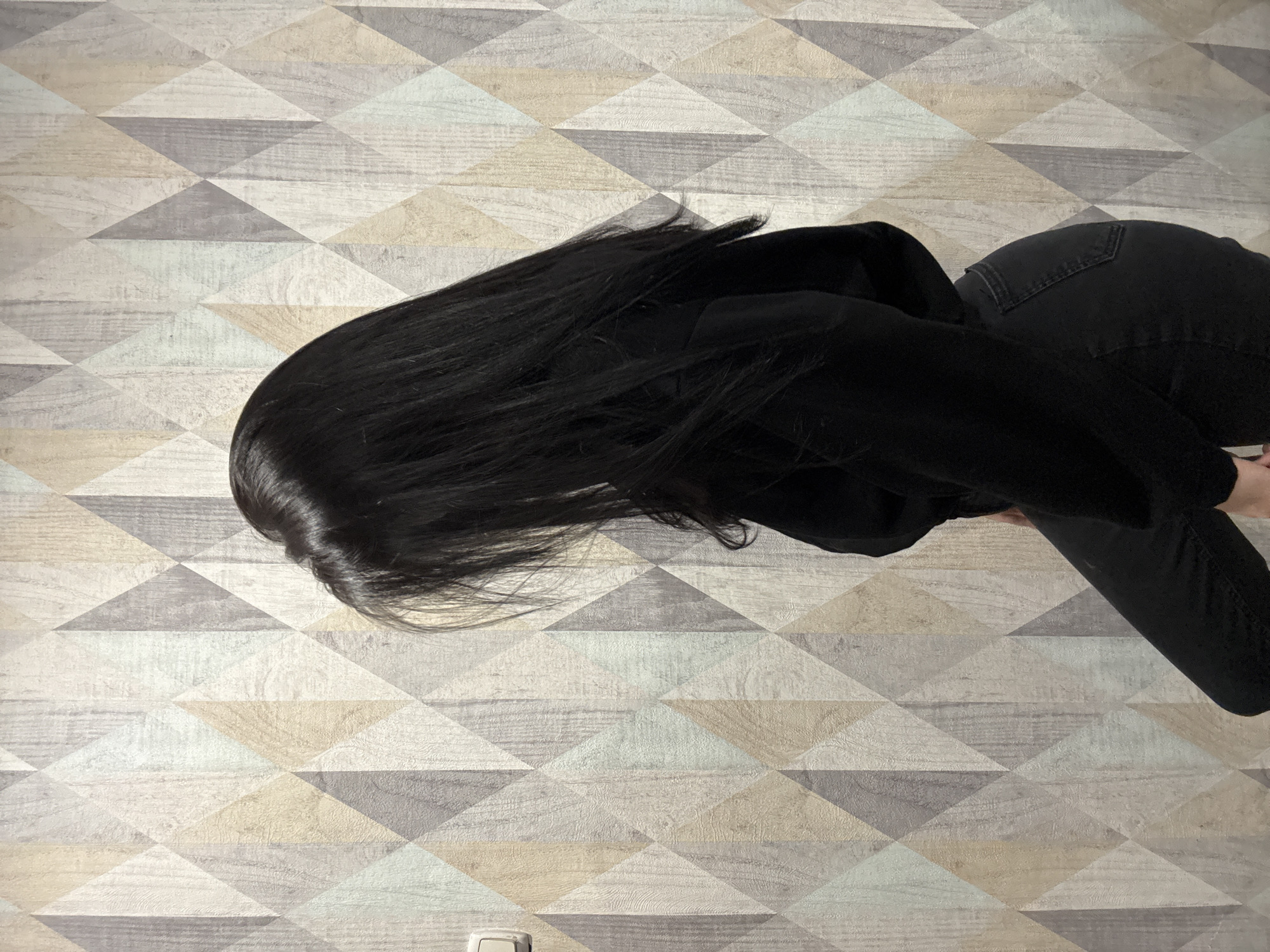A top-down view depicts a person lying on a surface covered with a geometric pattern of interlocking triangles in varying shades of grey and beige. The person has long, black hair that is spread out on the surface. They are wearing black clothing, including what appears to be a hooded top and jeans. The location is not specified but the presence of the city suggests that this is an indoor setting.