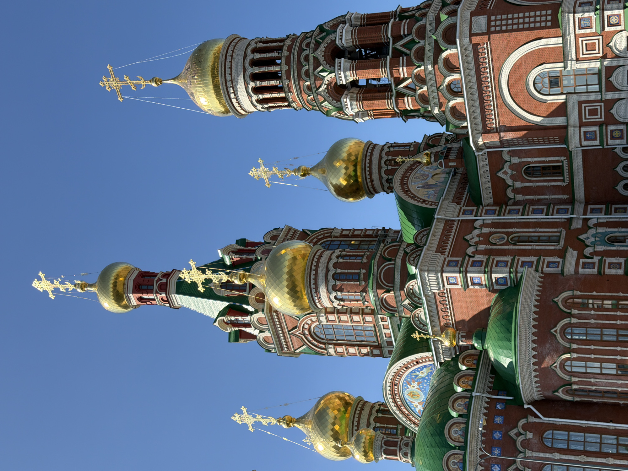 This is a medium shot, eye-level, outdoor photograph of the red brick facade of a large, ornate Russian Orthodox church with multiple green and gold domes, and a tall bell tower. The sky is clear and blue. The domes are various sizes and shapes, many topped with golden crosses. The bell tower features open arches and decorative brickwork. The building is richly decorated with colored tiles and mosaic-like paintings above some of the windows. The sun is casting shadows on the building, indicating it is daytime. The image is sharp and clear, with excellent detail of the architectural elements. There are no people or other objects visible in the image.