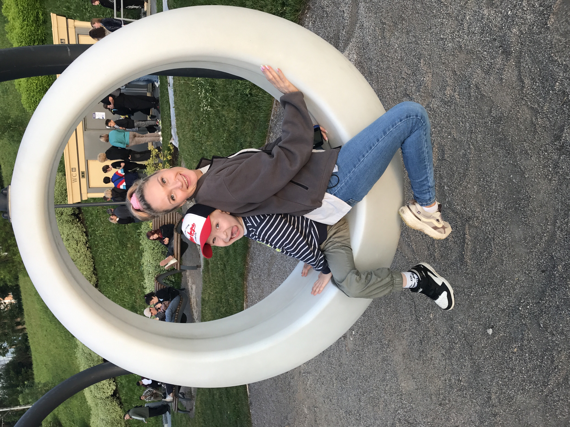 A woman and a young boy are sitting in a large, white, circular modern art sculpture in a park. The woman is on the right, facing forward and smiling at the camera. She has blonde hair pulled back in a ponytail and is wearing a grey jacket over a white shirt and blue jeans. The boy is on the left, facing the camera with his mouth open and sticking his tongue out. He is wearing a striped shirt, green pants, and a red and white baseball cap. In the background, there are other people walking and sitting, a building with "WC" signage, and a grassy hillside. The ground in the foreground is covered with gravel. The sculpture is located in Perm, Russia.
