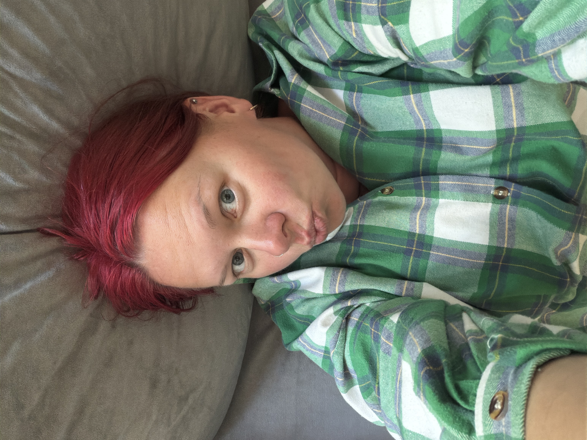 A woman with bright red hair is lying down, looking at the camera with her lips puckered and eyes wide. She is wearing a green and white plaid shirt. The background is a textured grey surface, possibly a couch or bed. The image appears to be a selfie, taken from a high angle.