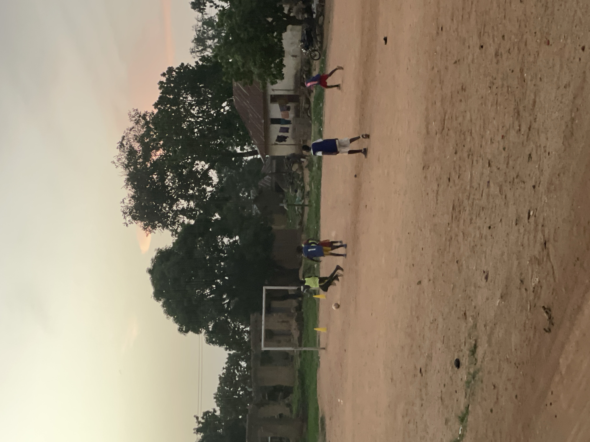 The scene depicts an outdoor soccer practice taking place in Barikin Gidar Kanat, Nigeria. Several young boys are engaged in the practice, one positioned as a goalkeeper in front of a makeshift goal composed of white metal posts, with two yellow cones placed on either side. A soccer ball is visible near the goal. Behind the goal, there is low green grass and a dilapidated building. Further back, a large tree with dense foliage dominates the left side of the image. Another building is visible with clothes hanging out to dry. To the right of this, there is a person bending over. The ground is dirt. The sky is overcast, and the lighting indicates that it is either early morning or late evening.