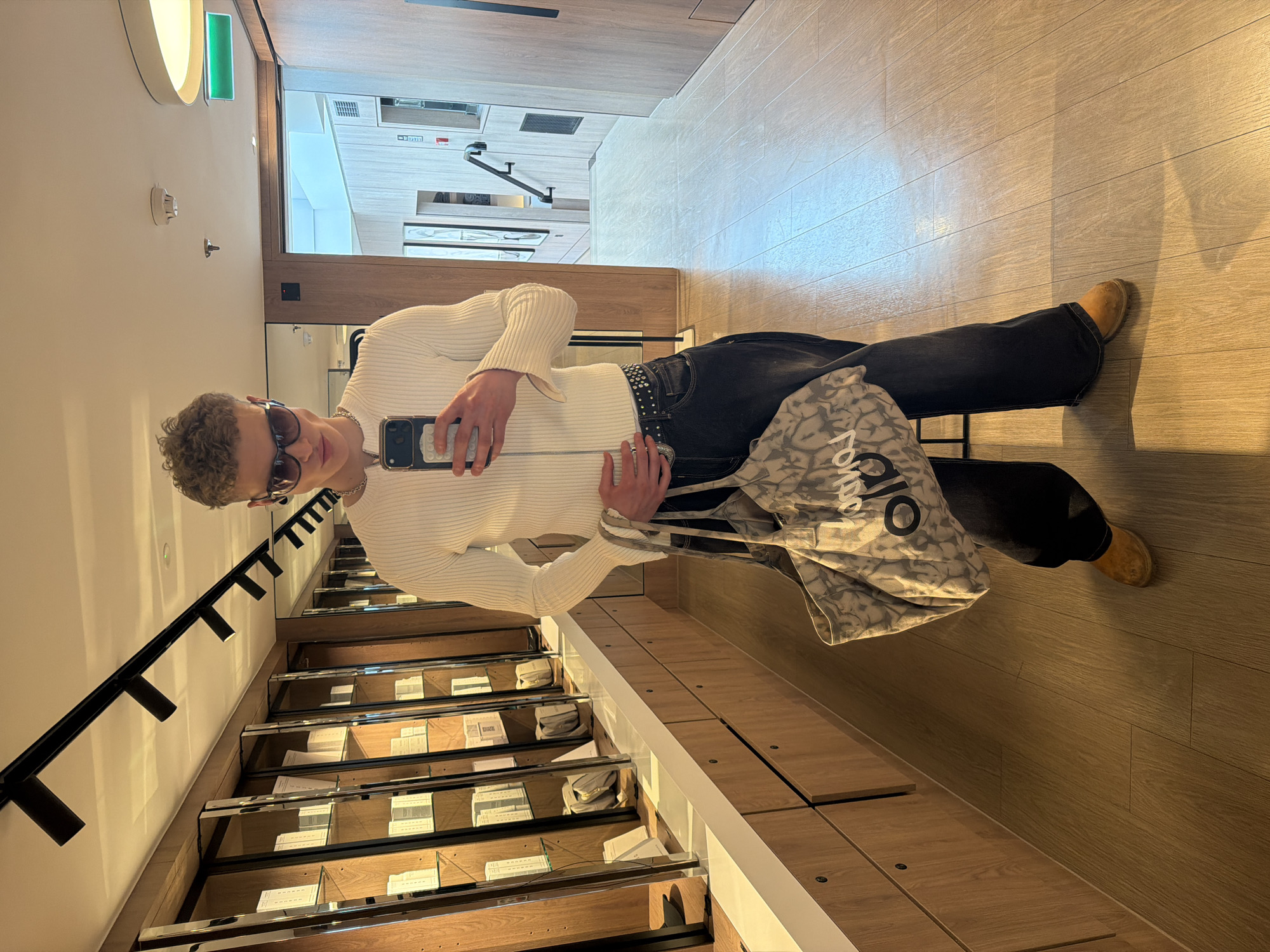 A full body image of a man standing in a retail store setting. The man has short, curly hair and is wearing sunglasses. He is wearing a white ribbed long-sleeve shirt, dark jeans with a studded belt, and brown shoes. He is holding a smartphone in his hand, presumably taking the photo. A gray patterned bag is also visible.
The store has a multi-level design with glass shelves displaying various items. Lighting fixtures are mounted on the wall and ceiling. The store appears to be modern and well-lit.
Based on the presence of "Alo" on the bag and retail setting, it's likely in London, United Kingdom. This is a standard photo of a person in a clothing retail setting.