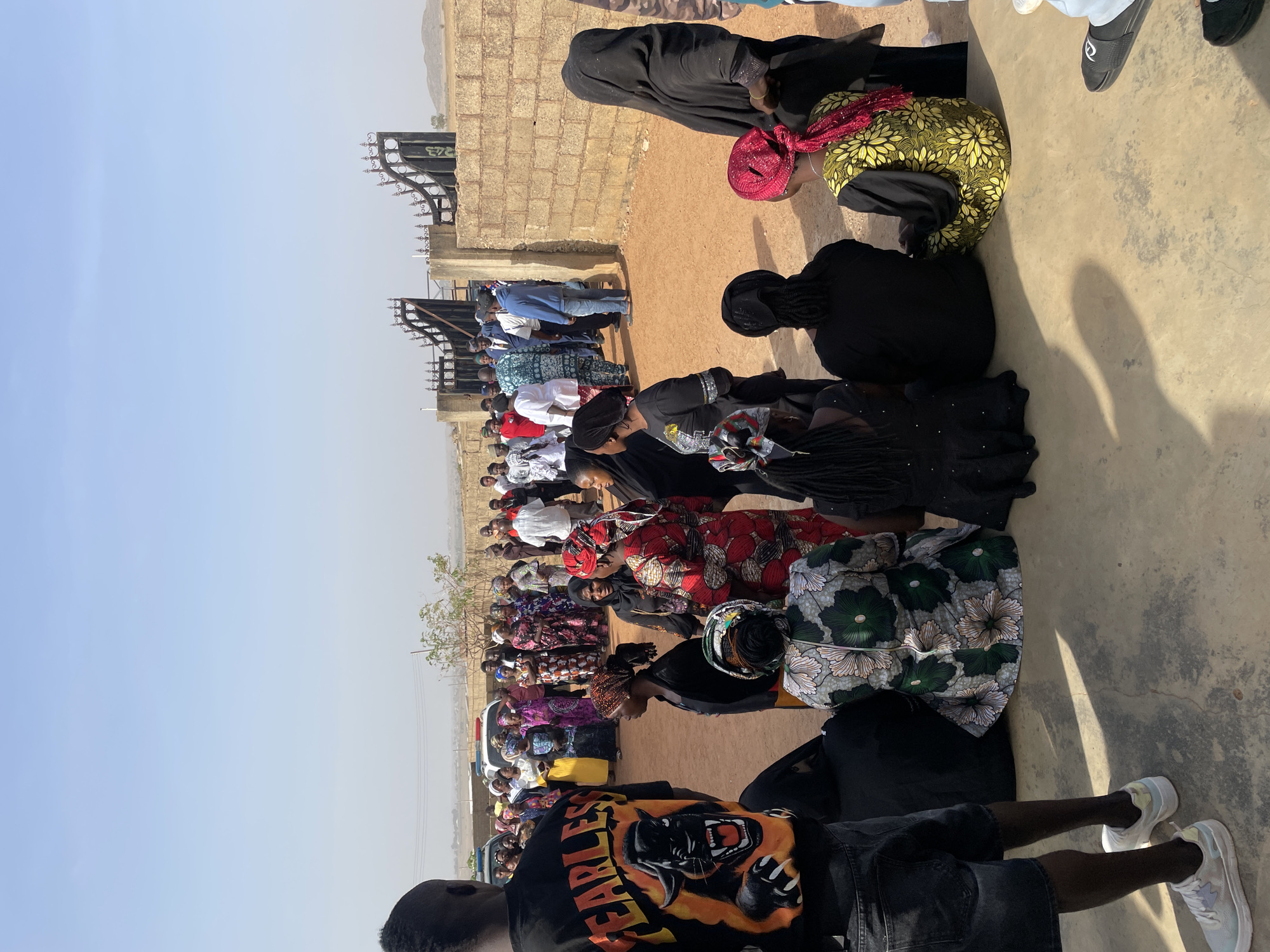 An outdoor gathering in Du, Nigeria, is captured under a bright, hazy sky, suggesting midday or early afternoon. The scene features a large group of people, primarily women, many of whom are kneeling or sitting on the concrete and dirt ground, facing towards the left of the frame. Most women are dressed in colorful, patterned traditional African wax print fabrics (Ankara) and head coverings, interspersed with some wearing dark, modest attire.
In the immediate foreground, the back of a person wearing a black t-shirt with a prominent "FEARLESS" graphic, black shorts, and white sneakers is visible. Further back, more attendees, including some men in Western clothing, are seen standing and sitting. The backdrop includes brick buildings and metal gate structures, with distant hills visible on the horizon. The collective posture of the crowd suggests an organized communal event, possibly a ceremony or religious gathering, characterized by a focused and orderly atmosphere.