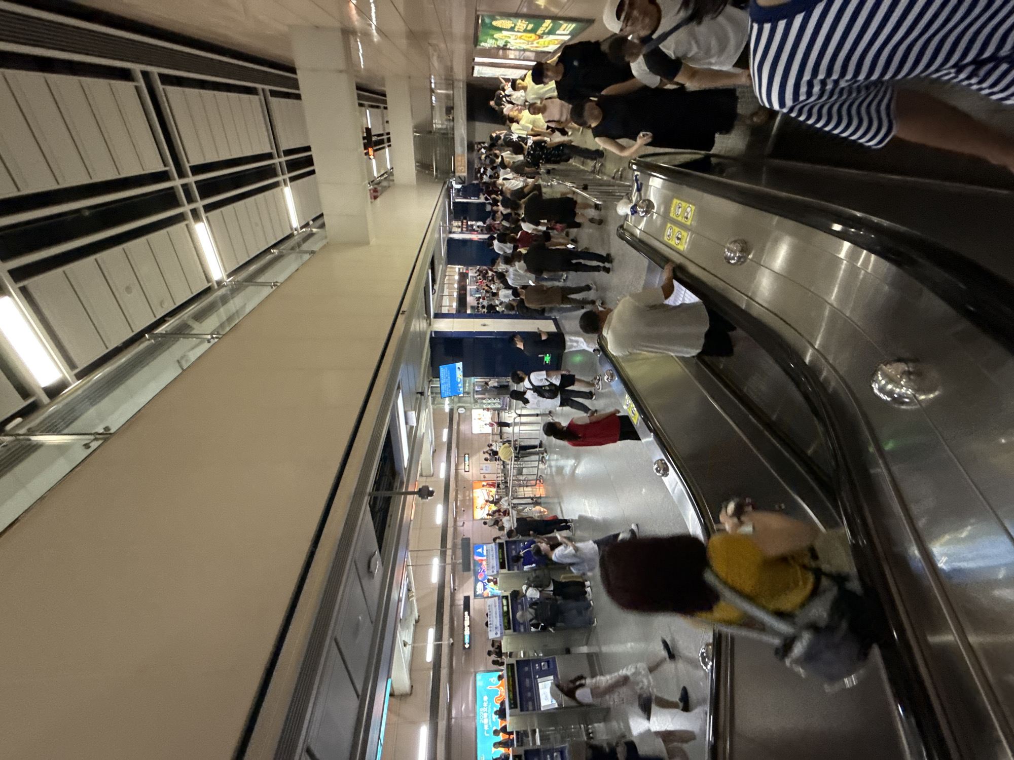 The image depicts a busy metro station escalator in Haizhu District, China. A diverse crowd of people are seen ascending and descending the escalators and walking through the station. The environment is well-lit with fluorescent lighting and features modern architecture, including metallic escalators, ticket machines, and signage. People are dressed in casual attire, carrying bags, and interacting in various ways, suggesting a typical commute or travel scenario. The overall scene conveys a sense of movement and daily activity within a public transportation hub.