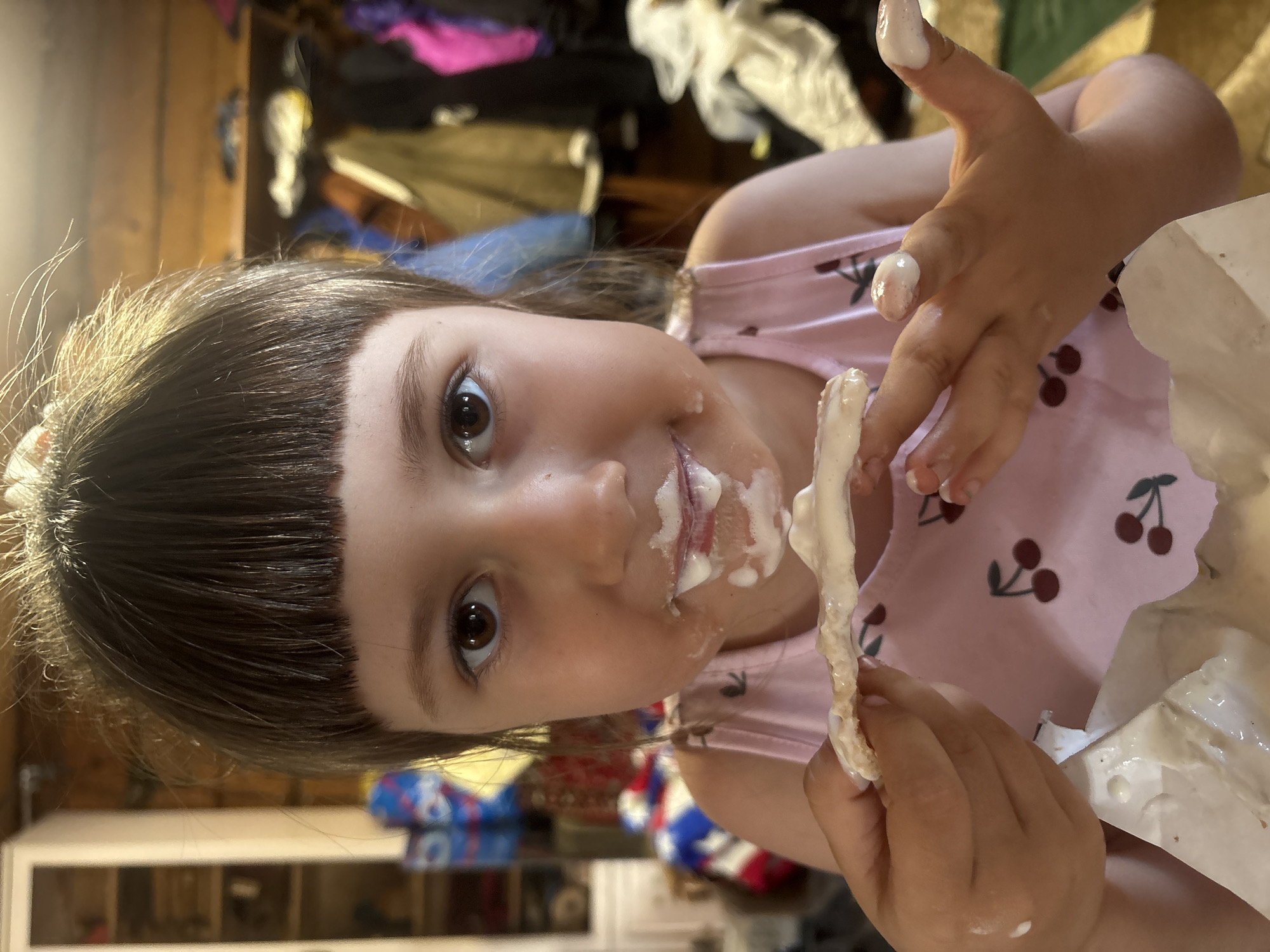 A young girl with dark hair and bangs is holding a food item covered in white cream. The cream is also smeared around her mouth and on her fingers. She's wearing a pink cherry-print shirt. The background is somewhat cluttered, with wooden surfaces and various objects visible. The specific location, Nizhnyaya Chermoda, Russia, cannot be confirmed from the visual data.