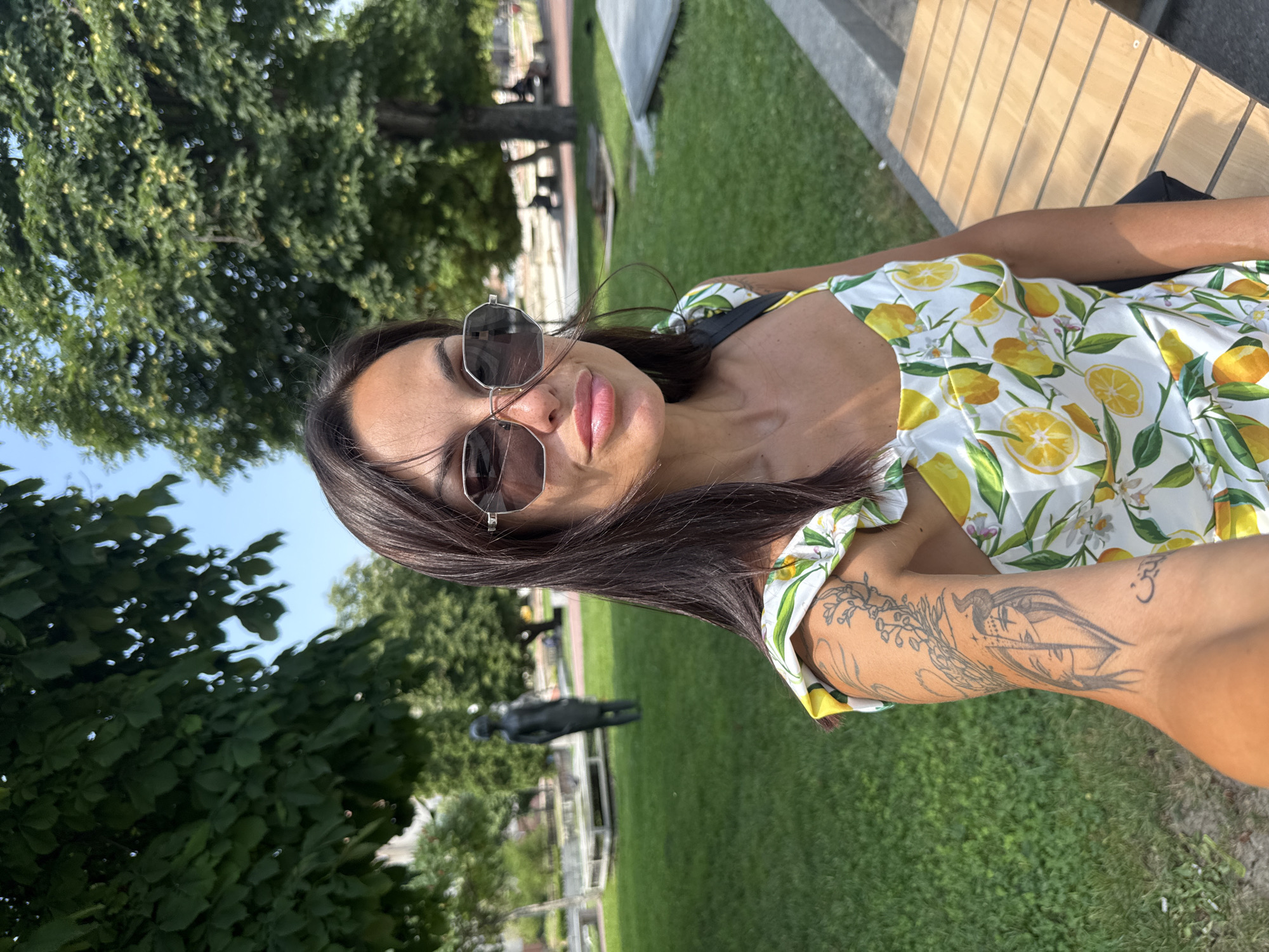 A woman is taking a selfie outdoors, as indicated by the sunlight and green grass. She is wearing a white dress with a lemon print and large sunglasses. Her left arm is visible, showing a tattoo of a face with horns and wings, as well as another smaller tattoo on her wrist. She is sitting on a wooden bench, and in the background, there are trees, a lawn, and a statue of a man in the distance. The sky is clear and blue. The overall mood appears relaxed and summery.