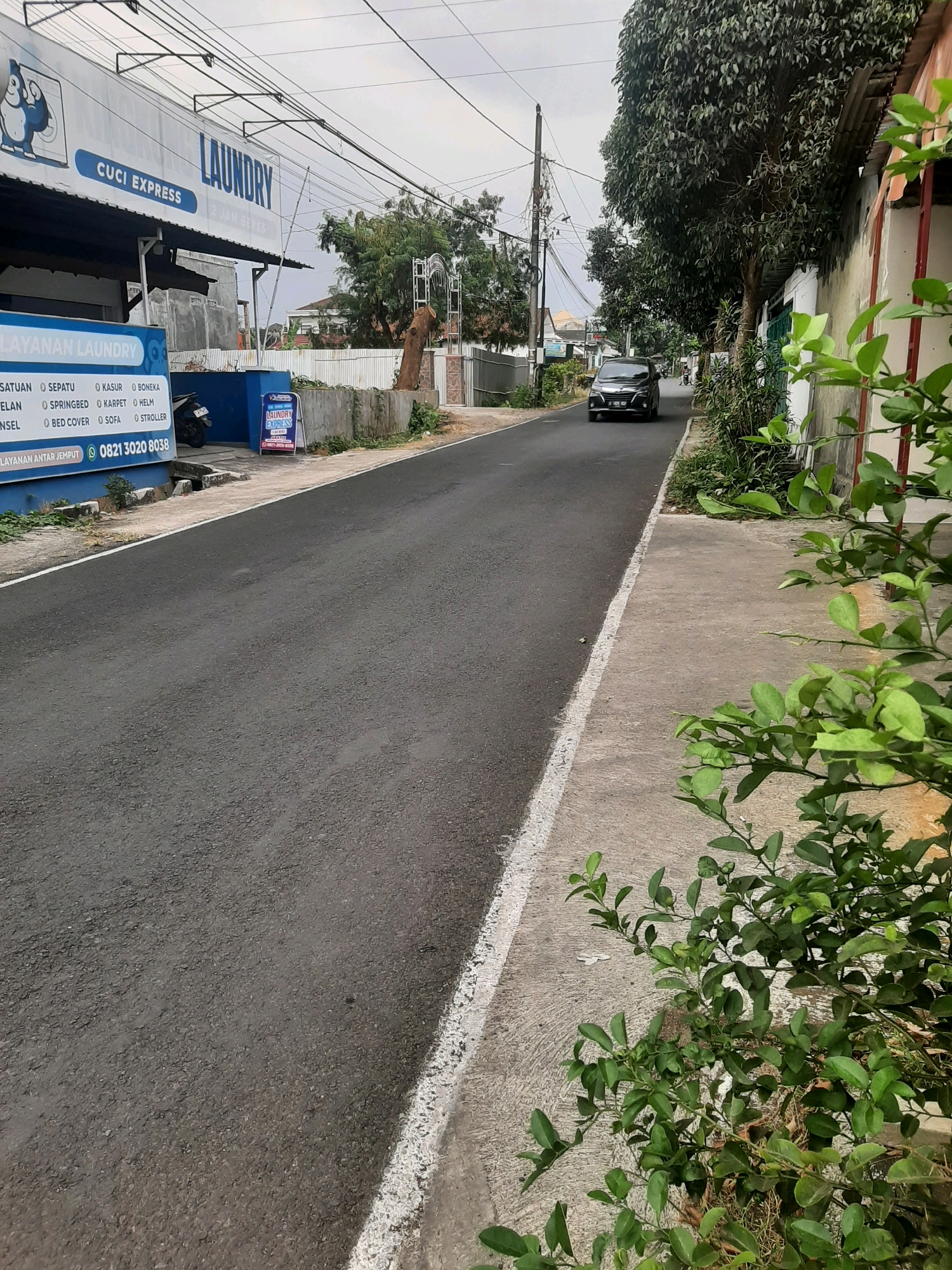 The image shows a street scene in Sumampir, Indonesia. On the left side of the road, a laundry business called "CUCI EXPRESS LAUNDRY" is visible, with a sign listing various laundry services. A black car is driving away from the camera down the street. The street is paved and has a white line marking the edge. There are trees and buildings lining the right side of the road, and some green foliage is in the foreground. The sky is overcast.