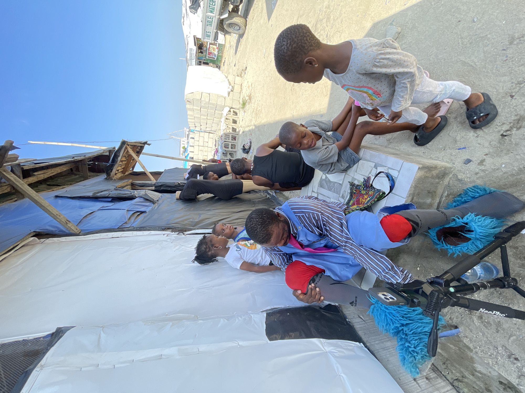 The image depicts an outdoor scene in what appears to be a temporary settlement or market area. In the foreground, a man wearing a blue striped shirt and gray leggings sits on a step, with a bright red covering on his left knee. His right leg is also covered in gray leggings. He is looking down at his hands, which are resting on his lap. To his left, a tripod with a phone mounted on it is visible. On his right, two children are seated on the ground. One child, a girl in a light gray long-sleeved shirt and white pants, stands looking towards the man, wearing large black sandals. The other child, a boy in a gray t-shirt and dark shorts, is seated on the step, looking downwards.
Behind the man, on a raised platform, two young girls stand. One girl wears a white t-shirt with a graphic print and has her hair in a ponytail. The other girl is partially visible. Further back, a group of people, including a woman in a black top, are standing or sitting near what appears to be a temporary structure made of white sheeting and dark blue tarpaulin, supported by wooden beams.
In the background, concrete blocks are stacked. A white truck with "NK'S TABLE WATER" printed on its side is visible to the right. The ground is sandy or dusty. The sky is clear and blue. The setting suggests a community that has experienced displacement or is living in challenging conditions, possibly in a developing urban or peri-urban area. The presence of mannequins near the temporary structure suggests a commercial activity. The location is identified as Iranla, Nigeria.