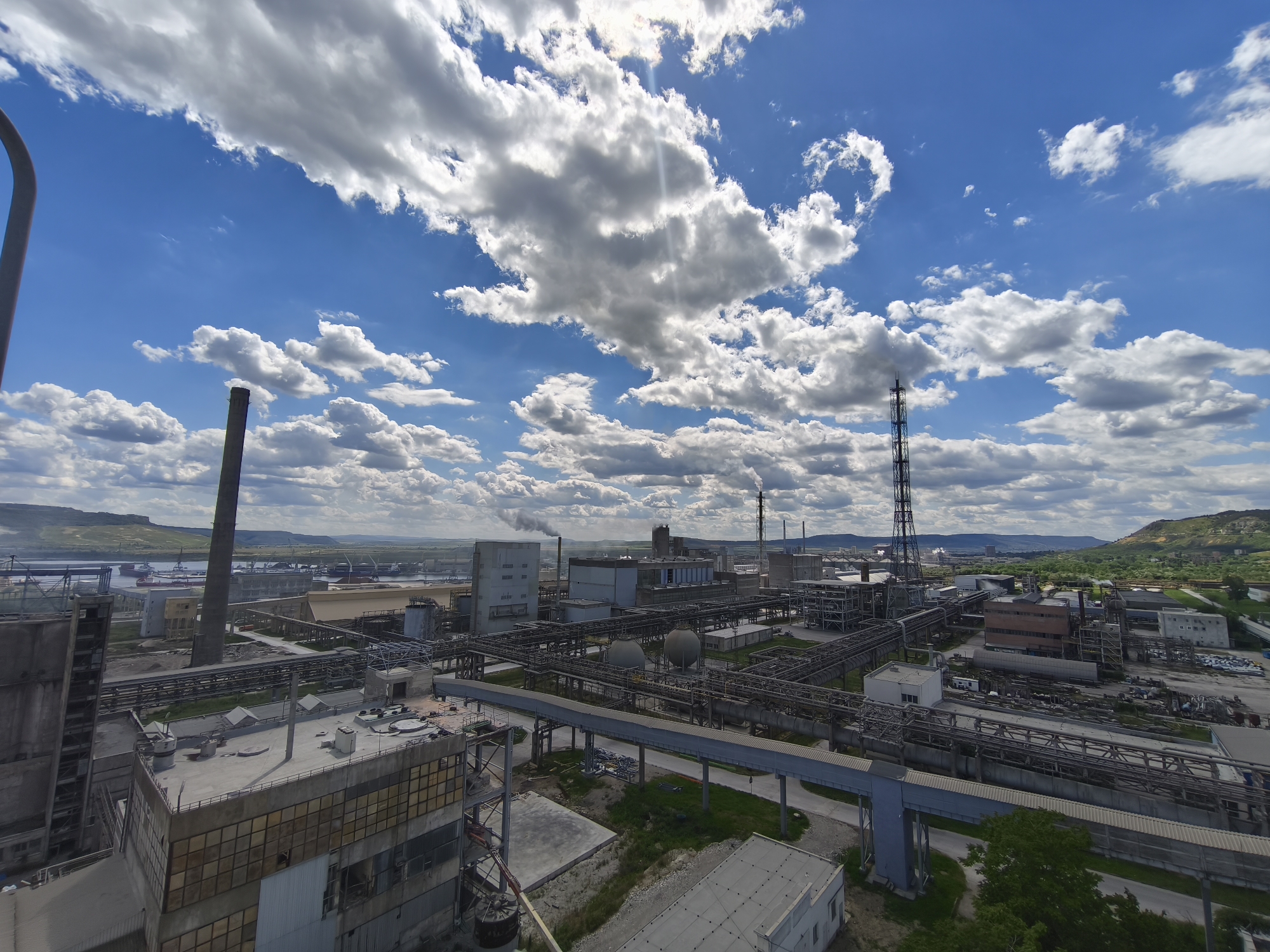The image shows an industrial complex under a sky with scattered clouds. The location is Unknown, Unknown. The scene includes multiple structures such as factory buildings, smokestacks, and an intricate network of pipes and walkways. Some smoke emanates from one of the smokestacks. In the distance, a body of water is visible along with hills.