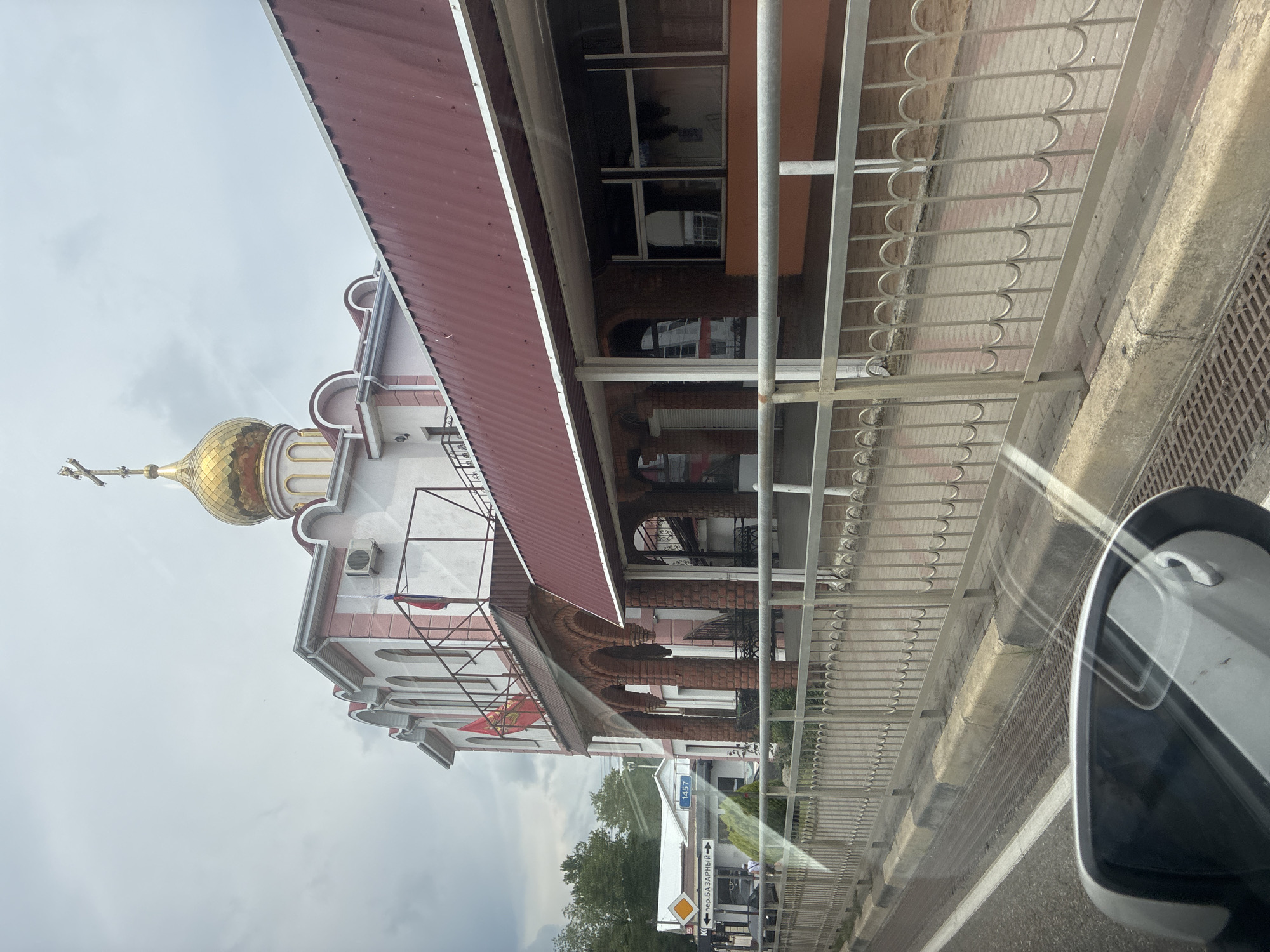 The image shows a building with a gold dome and a cross on top, characteristic of an Orthodox church, located in Arkhipo-Osipovka, Russia. The building has white walls with pink trim and arched windows. A red metal roof extends from the main building. There is a metal fence in the foreground, separating the sidewalk and a road. Several Russian flags are visible on the building. To the left of the church, there are signs, one of which reads "пер. БАЗАРНЫЙ", indicating a street name. The sky is overcast. The image appears to have been taken from inside a vehicle, as evidenced by the reflection in a car's side mirror in the lower left corner.
