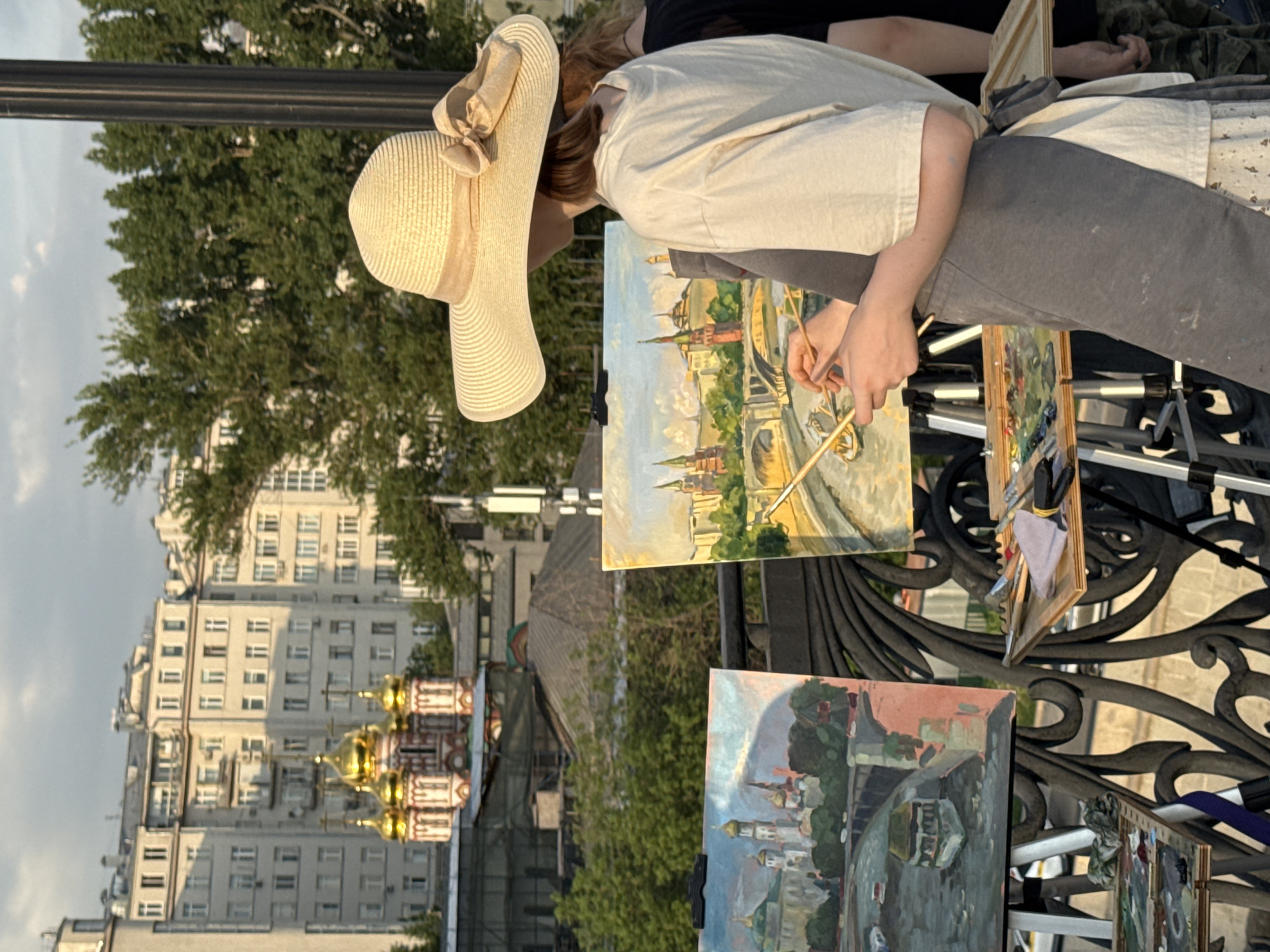 A person in a sun hat is painting a view of the Moscow Kremlin on an easel at an outdoor art display, located on a bridge in Moscow, Russia. The Cathedral of Basil the Blessed, with its distinct golden domes, is visible in the background, along with other buildings. The person is dressed in a light-colored shirt and gray pants. Other paintings are displayed on the bridge railing.