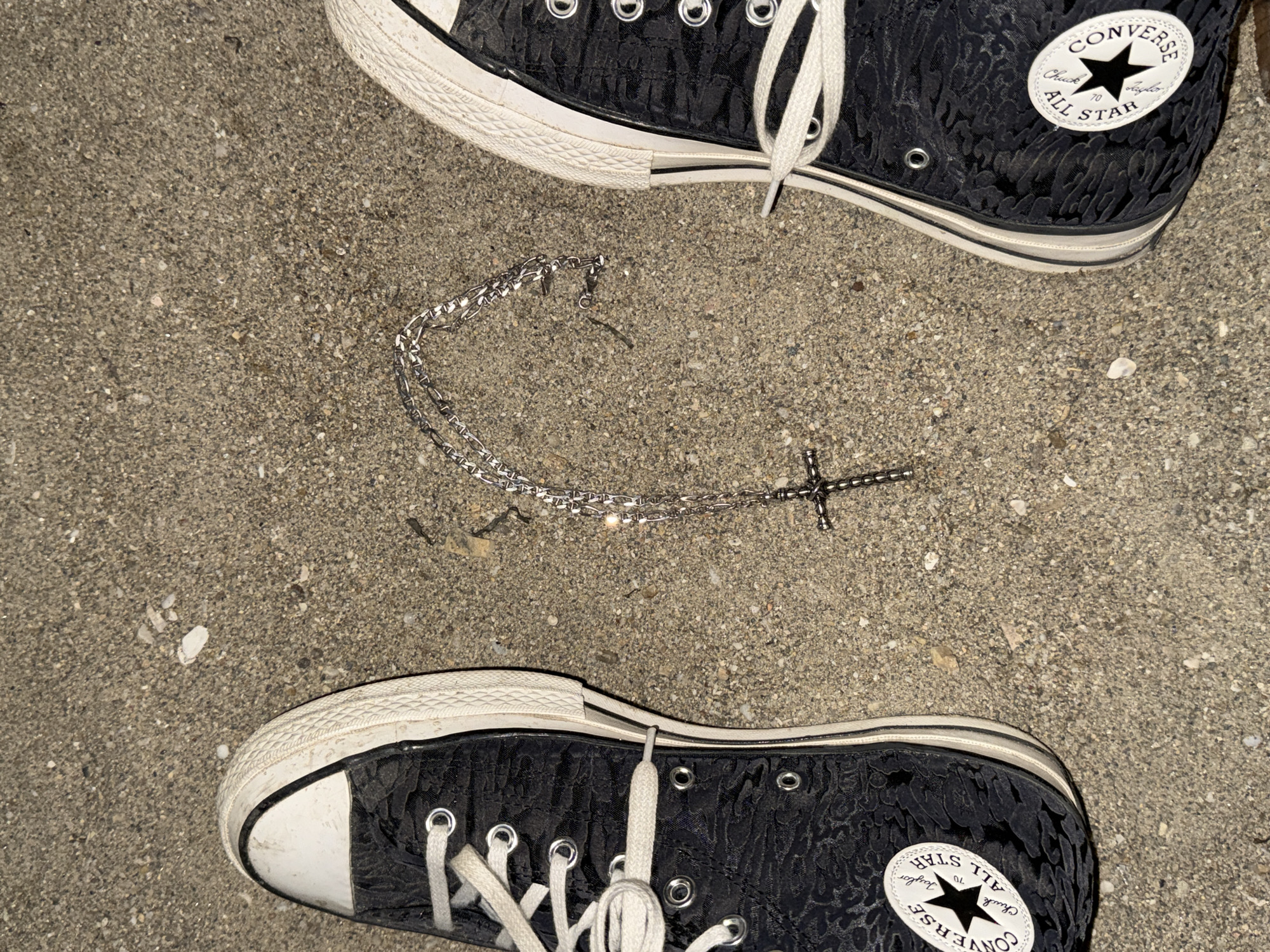 The image shows a pair of black Converse All-Star sneakers on a sandy, gravelly surface, likely a beach or a path in Nea Karvali, Greece. A silver chain with a cross pendant is lying on the ground between the shoes. The sneakers are placed with their toes pointing outwards. The surface is a mix of sand and small stones, with a few white specks scattered around. The lighting suggests it is daytime, possibly late afternoon or early evening given the shadows. No people are visible in the image. The scene is static and does not depict any action or interaction.