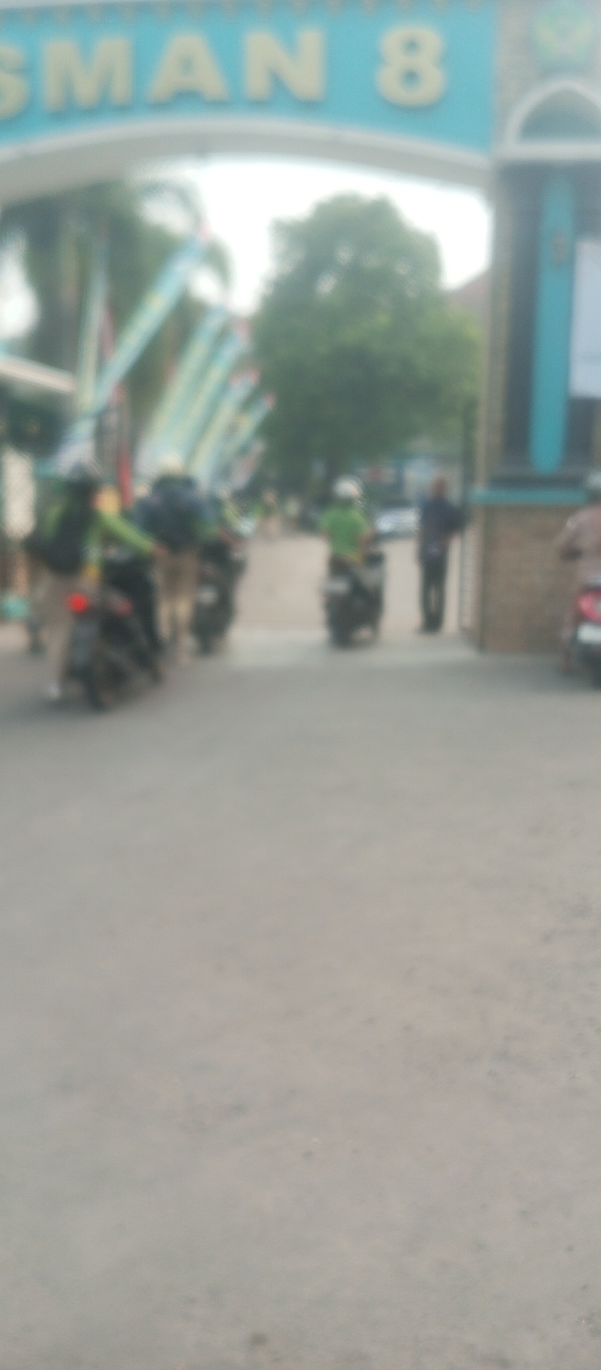 In front of the entrance to SMAN 8 Kediri City, Indonesia, several individuals on motorcycles are gathered. The sign above the entrance reads "SMAN 8". The scene appears to be an outdoor setting, with a paved road and some foliage visible in the background. The people and their motorcycles are mostly blurred, suggesting movement or a low-resolution image. Some individuals are wearing helmets, and their clothing suggests a casual or school attire. The overall atmosphere is that of a typical school entrance on a normal day, possibly during student arrival or departure.